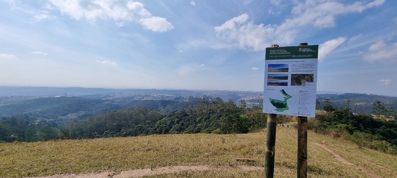 Pico do Votussununga – Morro do Cruzeiro. Um pico, céu azul sem muitas nuvens, dia ensolarado, grama verde bem baixa. Placa indicando o local