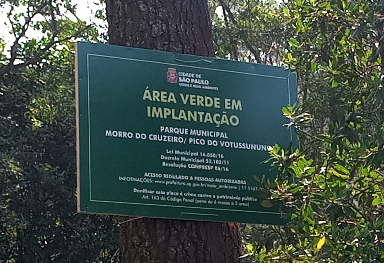 Placa verde com escritos "área verde em implantação". Atrás da placa, folhas de árvores