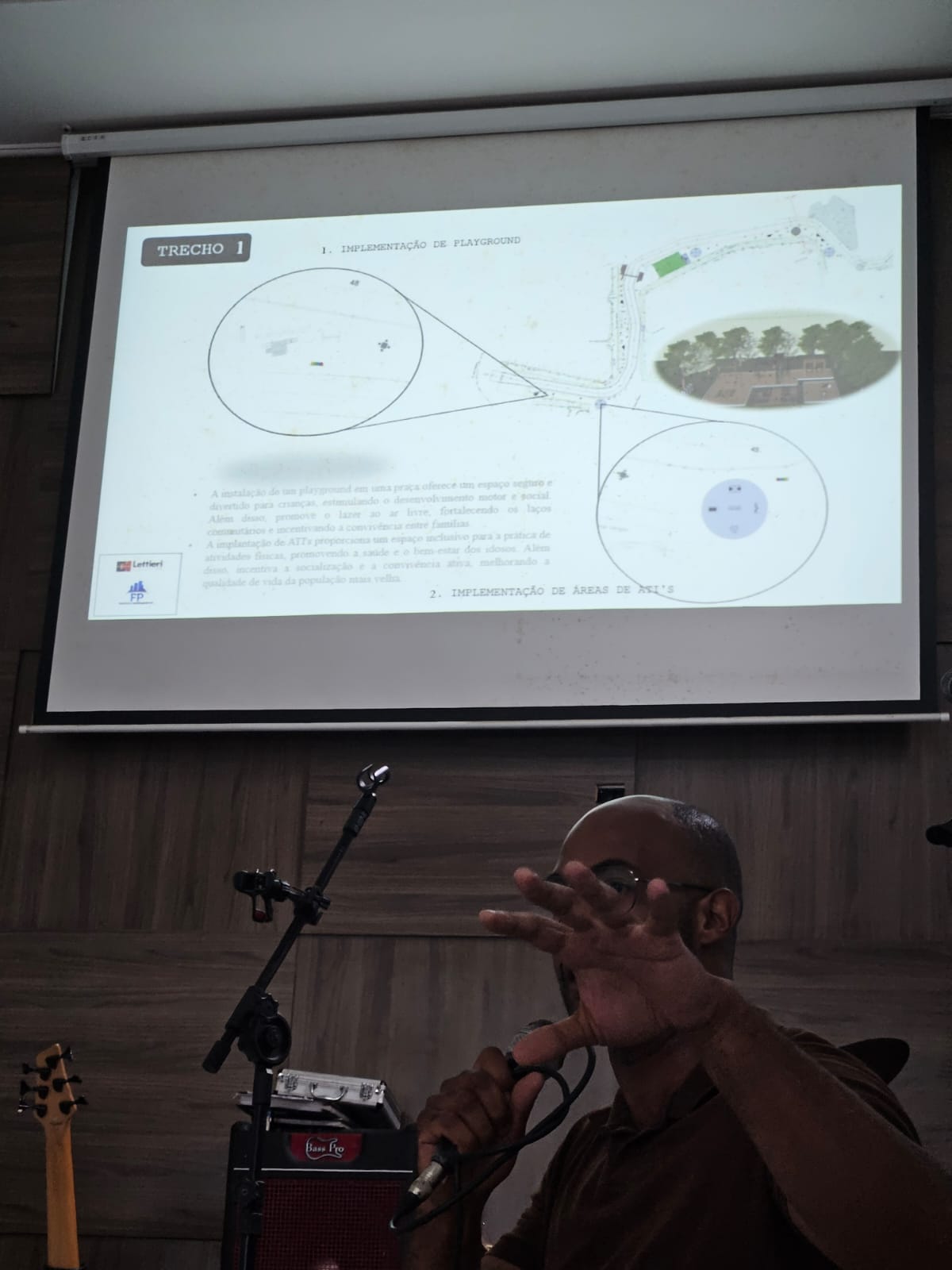 Homem, negro, careca, usando camiseta preta, com microfone na mão. Ao fundo, uma imagem por retroprojetor. 