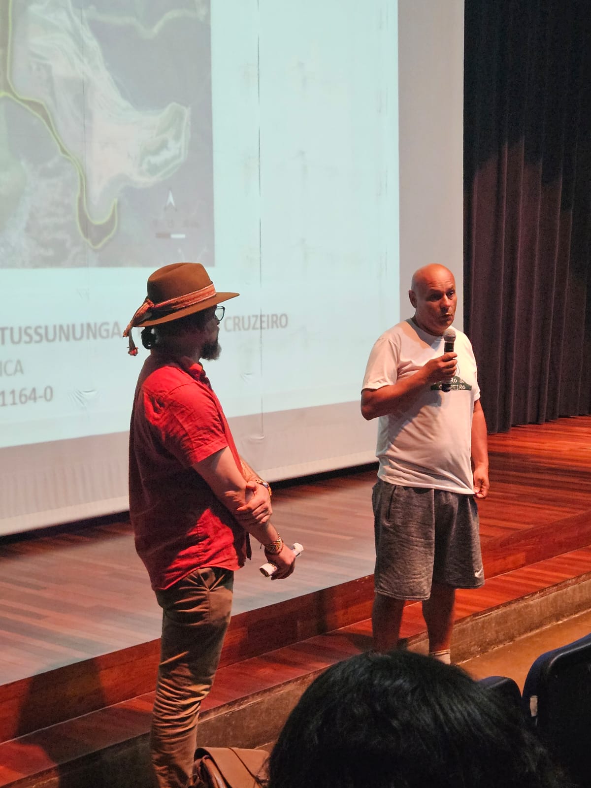 Dois homens na foto, o mais próximo usa camiseta vermelha, óculos e chapéu. O segundo, é careca e usa camiseta branca, está também com um microfone na mão