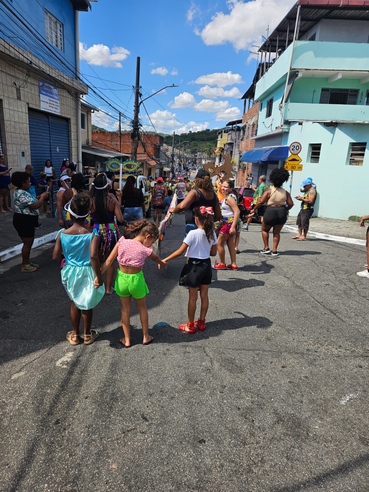 Pessoas na rua, em foco três meninas crianças de costas. Uma usa fantasia azul, outra veste blusa rosa e short verde, a terceira usa blusa branca e short verde. Um muro azul/verde água também aparece no fundo lateral direito da foto.