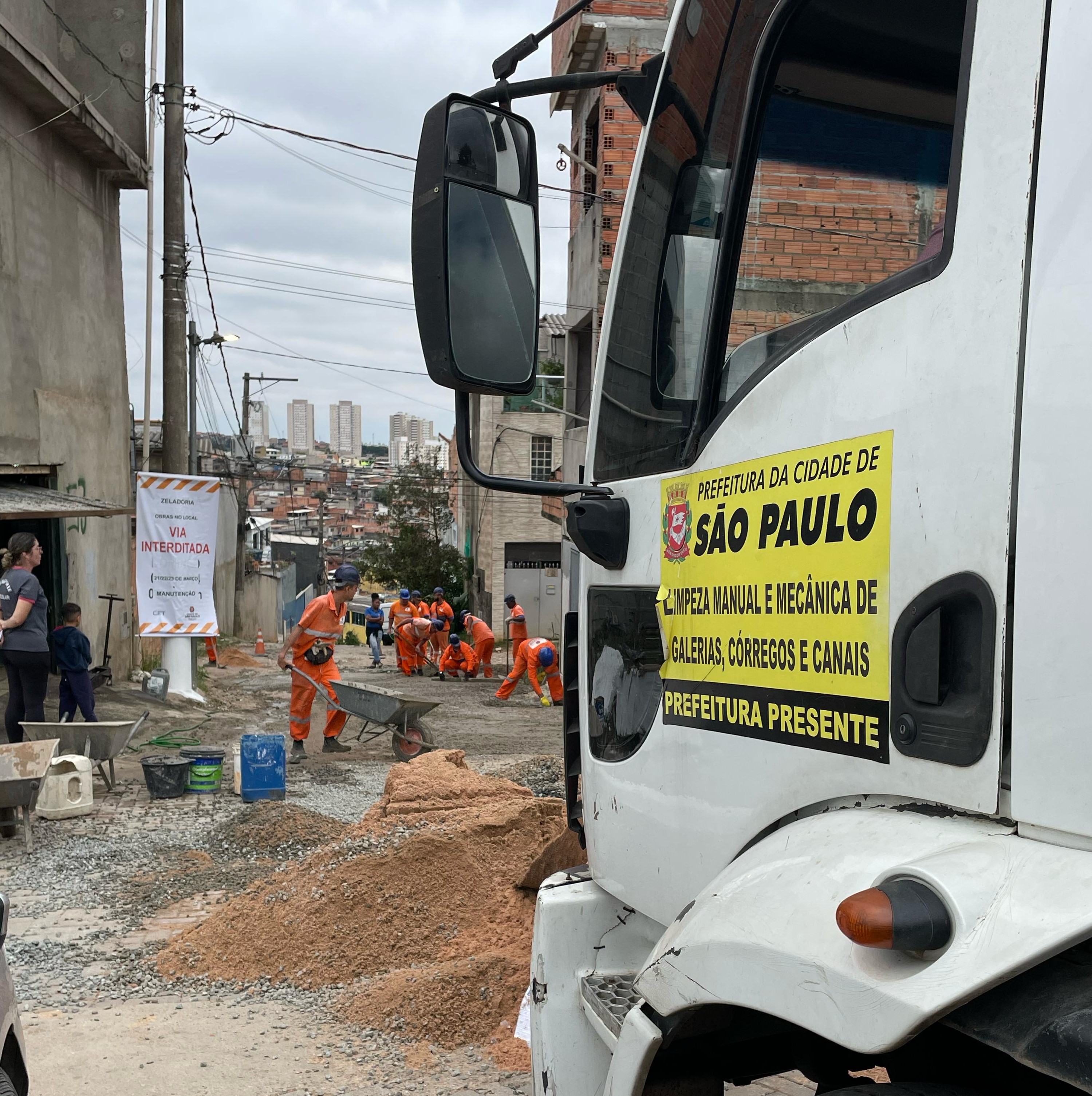 trabalhadores de laranja trabalhando em uma obra na rua, enquanto um caminhao se mantem na lateral da imagem com o banner da subprefeitura escrito 'limpeza manual e mecanica de galerias, córregos e canais' 'prefeitura presente'