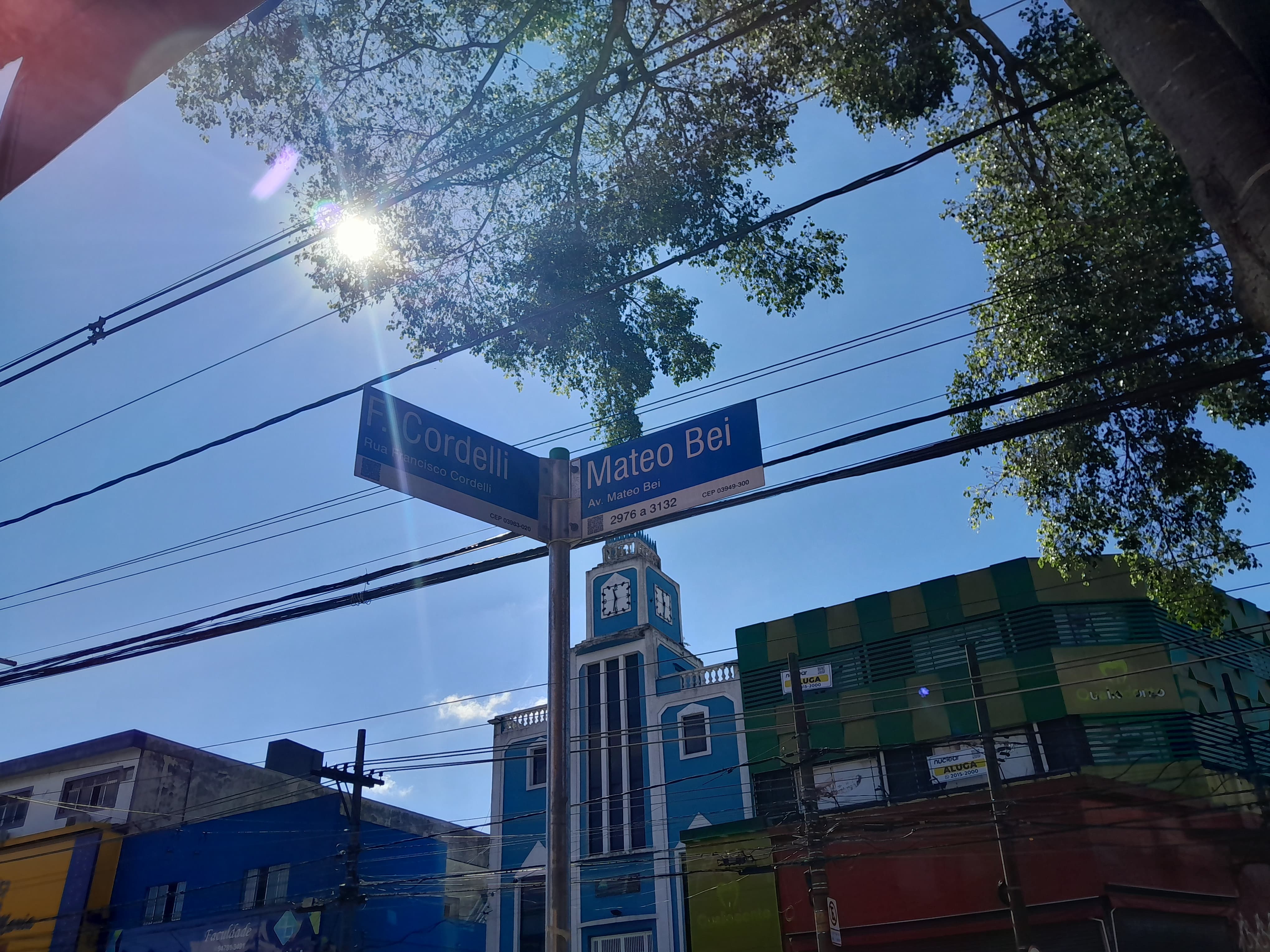 Ao fundo, céu azul com raios de sol iluminando a foto. Árvores aparecendo no canto superior direito da tela e, em foco da imagem, uma placa indicando a Rua Francisco Cordelli à esquerda e a Avenida Mateo Bei à direita.