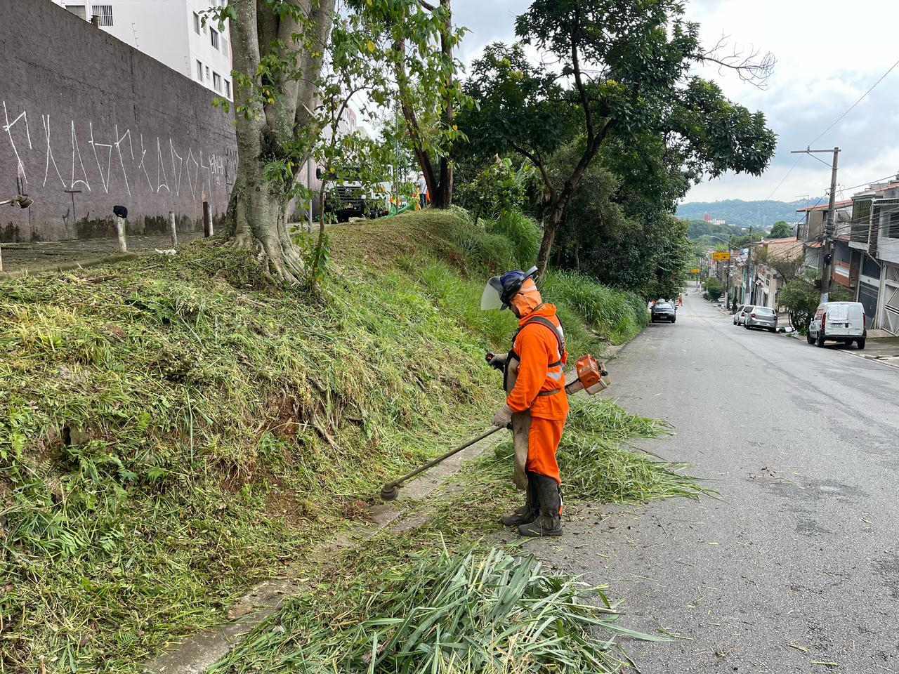 #PraCegoVer: Imagem mostra um funcionário da Prefeitura de São Paulo realizando um corte de mato em uma rua de São Mateus. Ele usa um EPI (Equipamento de Proteção Individual) e uniforme de cor laranja. 