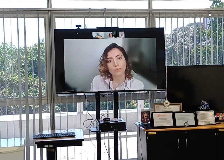 Pessoa participa de uma reunião por videoconferência exibida em uma tela de televisão em ambiente institucional da Subprefeitura de São Miguel Paulista. Ao redor da tela, há equipamentos de informática, certificados e materiais de escritório, indicando a realização de um encontro remoto para discussão e alinhamento de atividades.