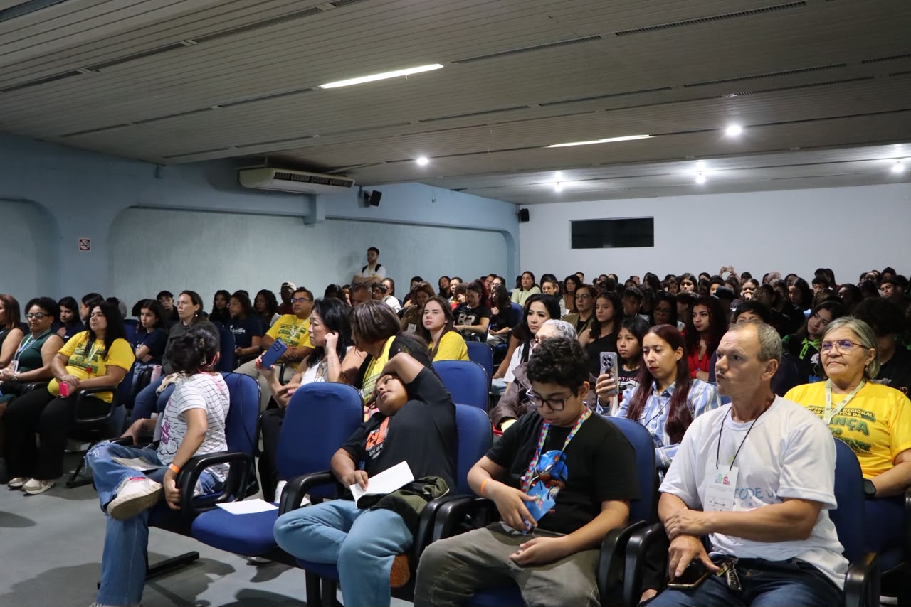 Público participa de um encontro realizado em um auditório, com cadeiras organizadas em fileiras e ampla presença de estudantes e moradores da região. A atividade reúne dezenas de participantes em um espaço fechado, voltado à troca de informações, orientação e participação comunitária.