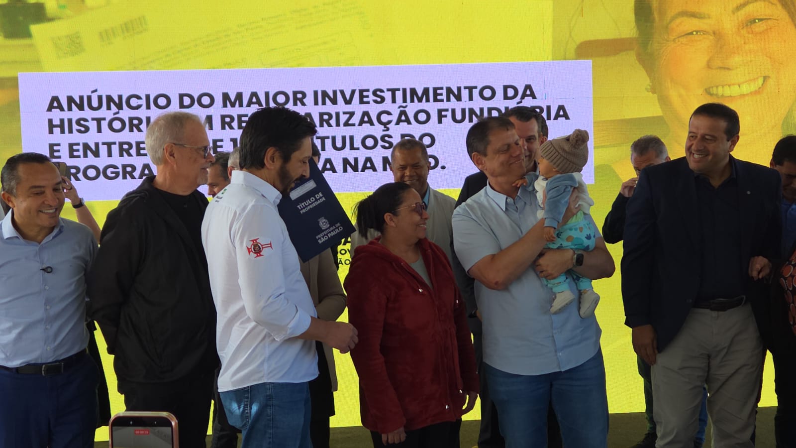 A imagem mostra um grupo de pessoas  reunido em um palco durante um evento público. Ao centro, um homem segura um bebê no colo e sua esposa está recebendo seu título de propriedade. Outras pessoas estão ao redor, todas de pé, participando do momento. Ao fundo, há um grande painel amarelo com texto sobre anúncio de investimentos e regularização fundiária