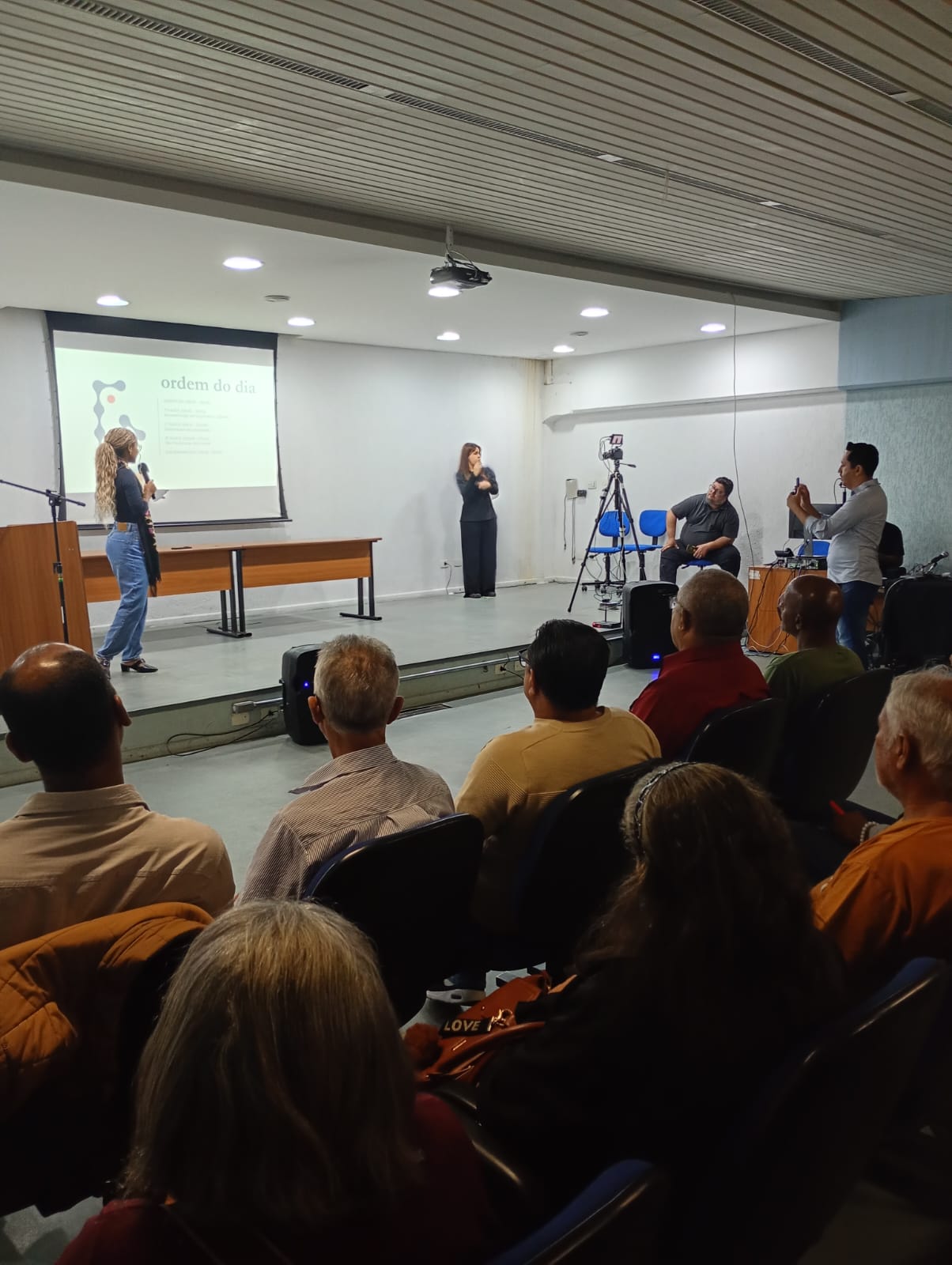 A imagem mostra funcionários públicos apresentando projetos de Orçamento Público com munícipes assistindo.
