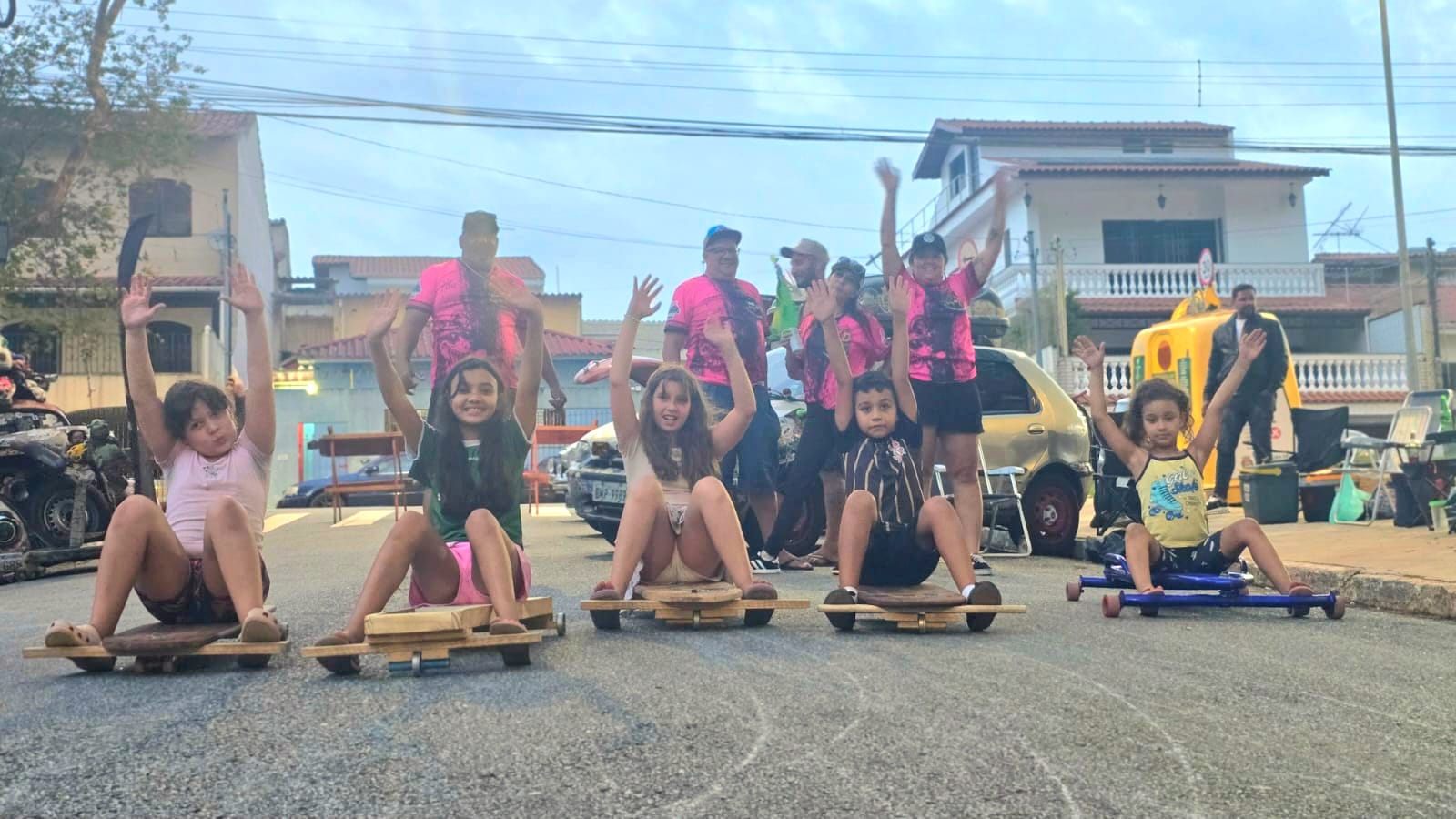 Crianças participam de uma atividade recreativa em via pública utilizando carrinhos de rolimã. Na imagem, os participantes estão alinhados na rua com os veículos artesanais, enquanto pessoas acompanham a atividade ao fundo, em um ambiente de lazer e integração comunitária.