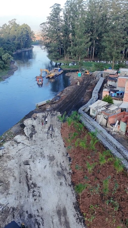 Área às margens do Rio Tietê passa por obras de recuperação ambiental e contenção. Na imagem, é possível observar máquinas operando próximo ao rio, uma embarcação de apoio e um muro de gabião instalado ao longo da margem, ao lado de residências. A intervenção integra ações de estabilização do terreno, proteção da margem e recuperação da área ribeirinha.