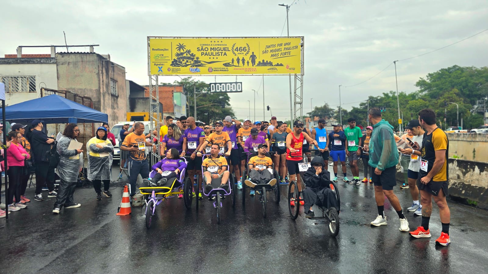 Participantes se reúnem na linha de largada de uma corrida e caminhada realizada em comemoração aos 466 anos de São Miguel Paulista. A imagem mostra corredores, cadeiras adaptadas para participação inclusiva e equipes de apoio posicionadas sob o pórtico do evento, reforçando a integração entre esporte, lazer e acessibilidade. Ao fundo, é possível ver o cronômetro da prova e a estrutura montada para receber os participantes.