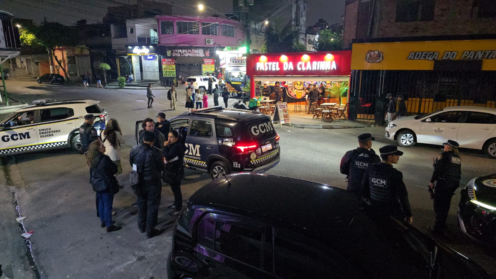 Equipes da Guarda Civil Metropolitana participam de ação de fiscalização noturna durante a Operação Adegas. Na imagem, viaturas e agentes estão posicionados em uma via comercial, em frente a estabelecimentos e pontos de alimentação, realizando atividades de monitoramento e ordenamento urbano na região.