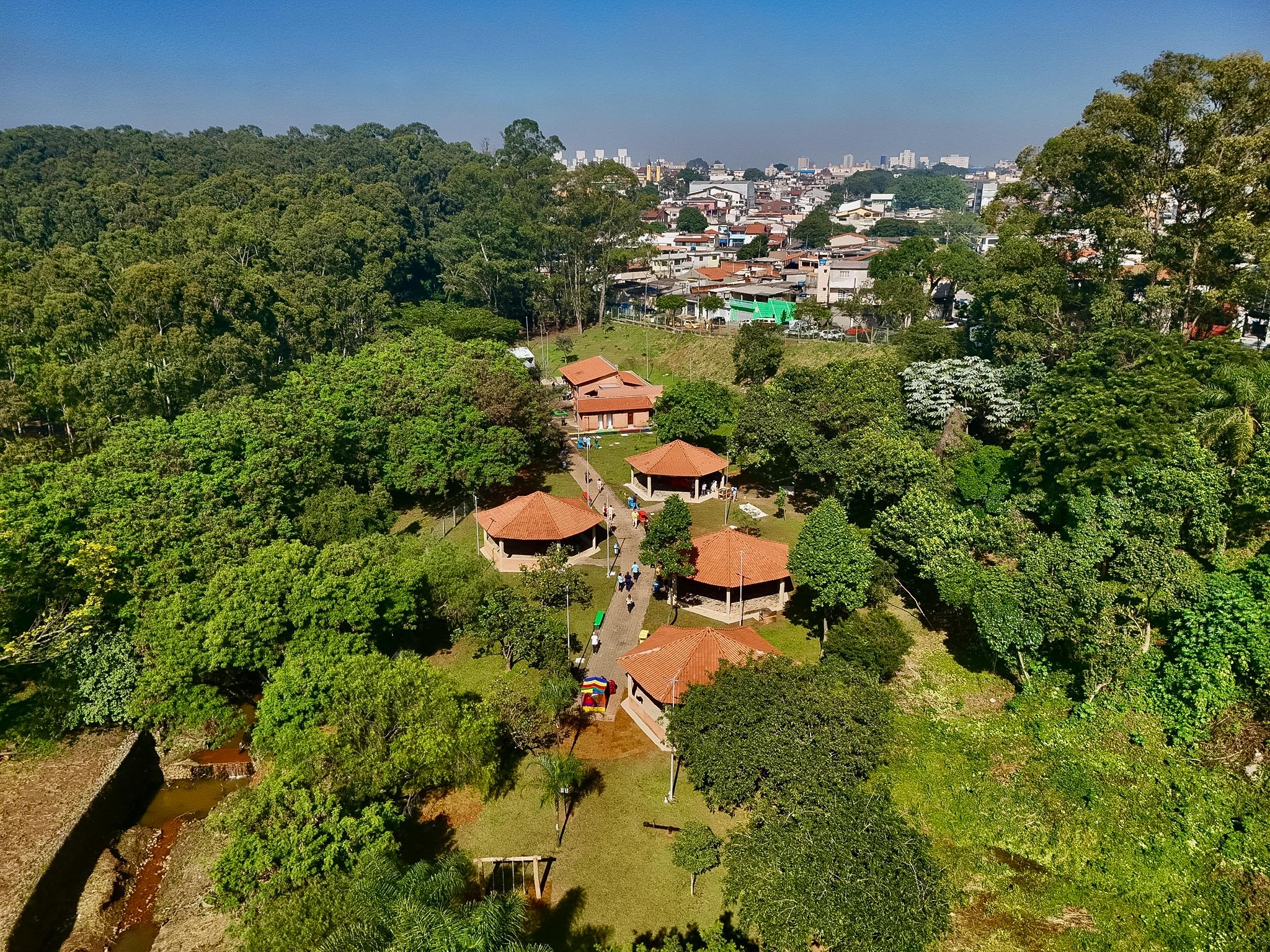 fotografia aérea que mostra a área de lazer ao ar livre do o parque, em um dia ensolarado. O local possui diversas construções com telhados de barro pessoas circulando por caminhos pavimentados.