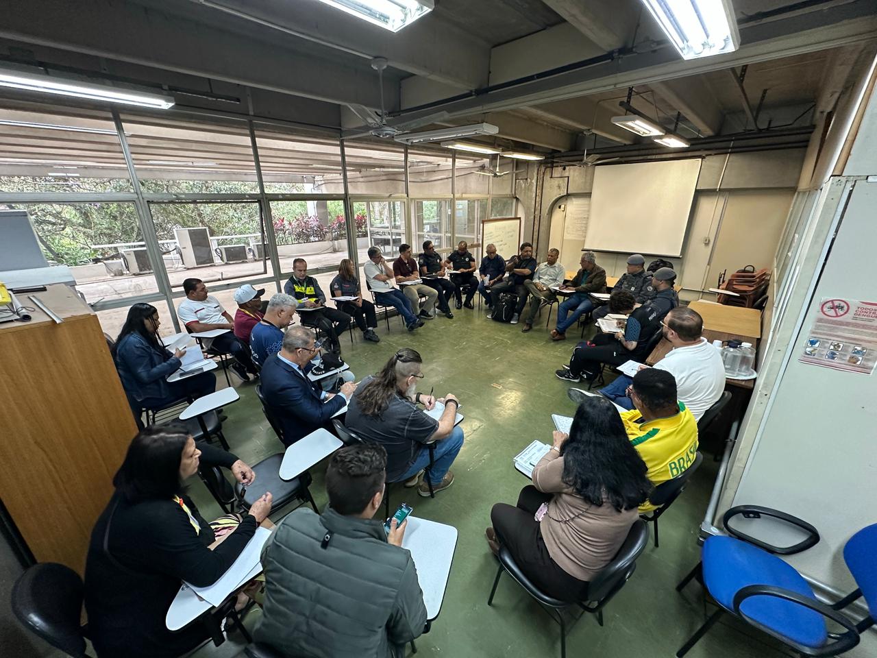 A imagem mostra um grupo de aproximadamente 20 pessoas sentadas em círculo em uma sala de reunião, participando da apresentação. 