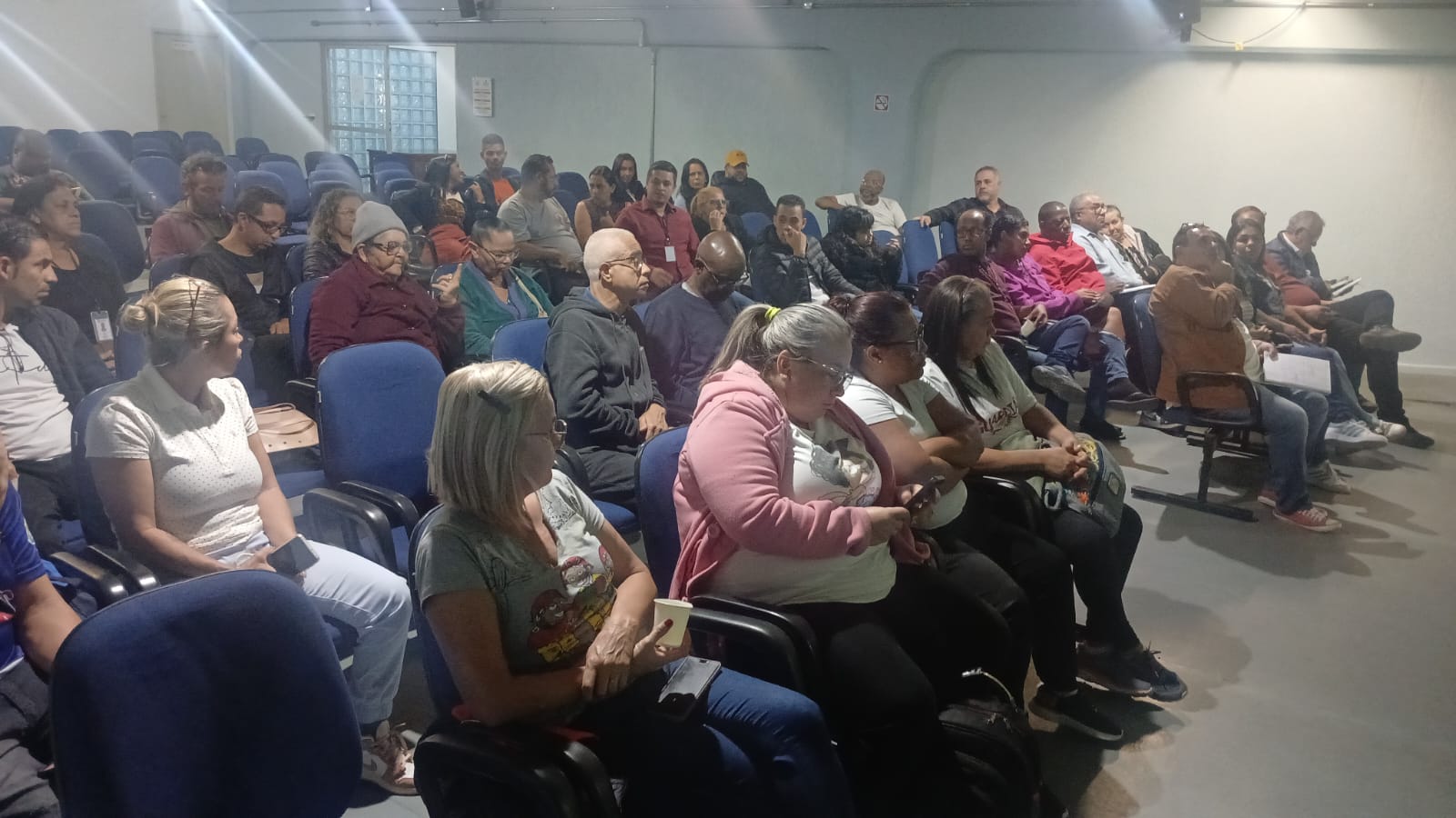 Pessoas participam de uma reunião em um auditório da Subprefeitura de São Miguel Paulista. O grupo está sentado em cadeiras azuis enquanto acompanha apresentações e orientações sobre programas sociais e serviços essenciais. O encontro reuniu lideranças comunitárias para fortalecer o acesso da população a iniciativas voltadas à melhoria da qualidade de vida na região.