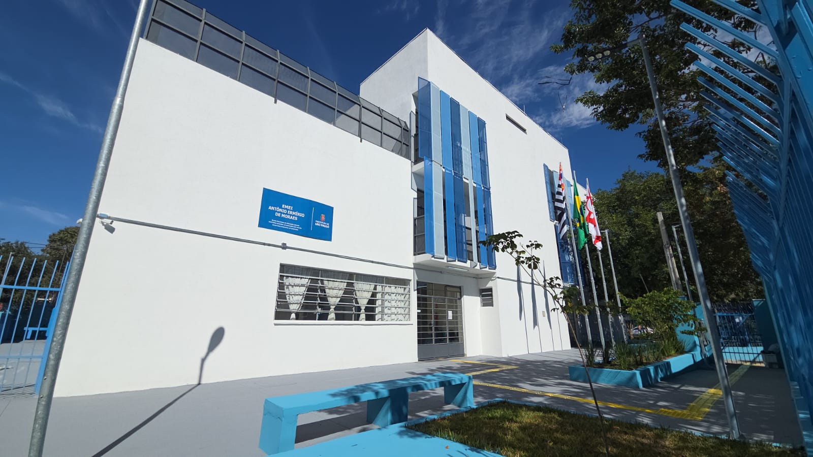 Fachada da EMEI Antônio Ermírio de Moraes, prédio moderno de dois andares com paredes brancas e detalhes em azul. A entrada conta com piso tátil, área ajardinada e mastros com as bandeiras do Brasil, do Estado e do Município de São Paulo, sob um céu azul.