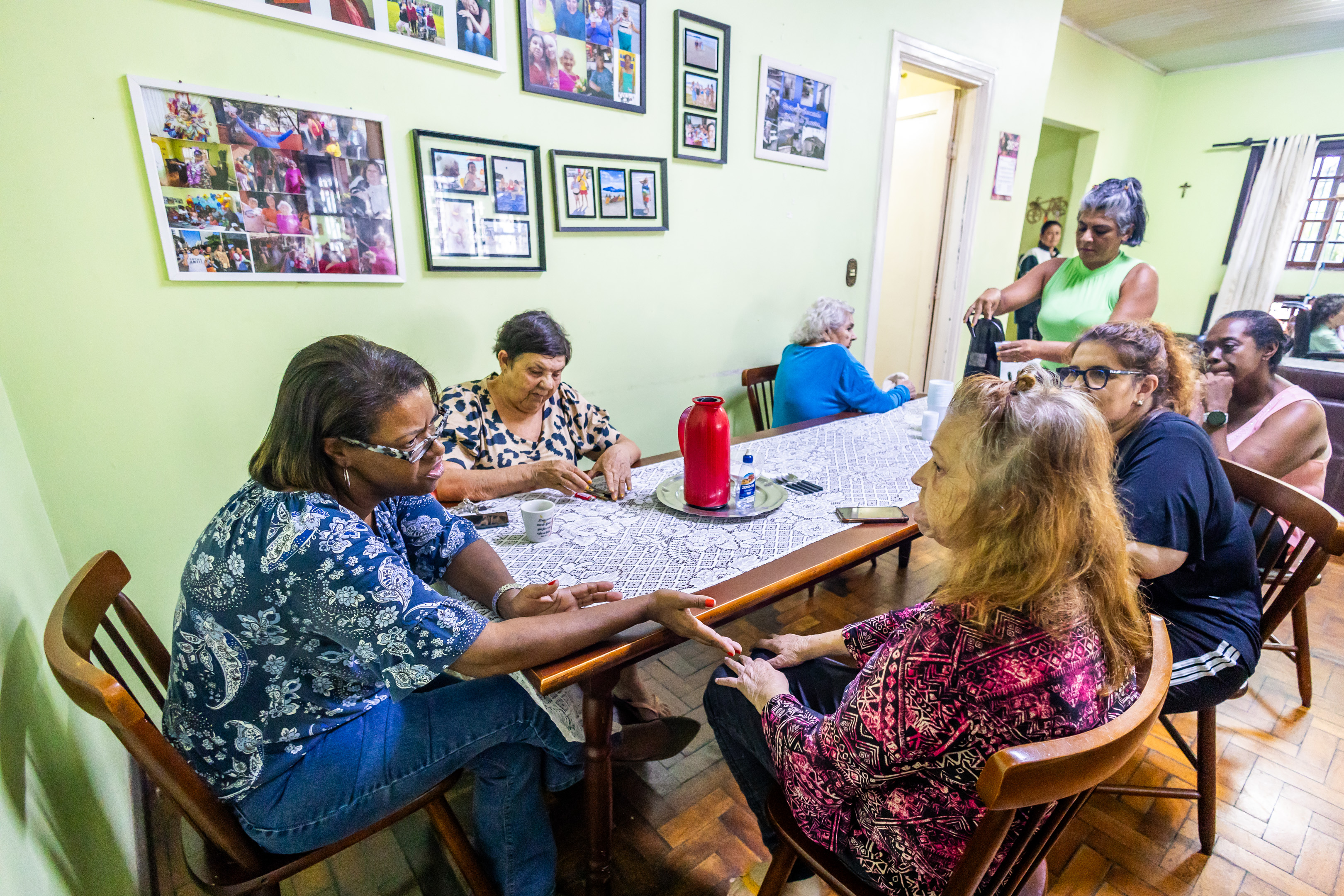 A imagem mostra um grupo de mulheres idosas reunidas em torno de uma mesa grande, dentro de uma residência com paredes claras e decoração afetiva. A mesa está coberta com uma toalha rendada branca e tem uma garrafa térmica vermelha, xícaras e outros objetos típicos de um momento de convivência, como celular, álcool gel e creme para as mãos.  Algumas mulheres conversam, enquanto uma delas parece estar fazendo um cuidado nas mãos de outra, sugerindo um momento de atenção ou carinho. Ao fundo, uma mulher de blusa verde serve bebida (provavelmente café ou chá) para outras senhoras, reforçando o clima de acolhimento.  Na parede, há diversas molduras com fotos de momentos vividos pelas moradoras ou participantes do local, o que cria uma atmosfera afetiva, familiar e cheia de memórias.  A cena transmite um ambiente de acolhimento, socialização e cuidado mútuo, típico de uma residência terapêutica ou espaço comunitário voltado para mulheres idosas, promovendo bem-estar e convivência ativa.