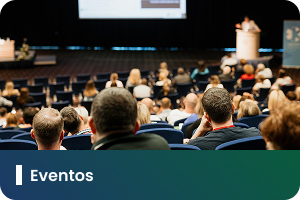 Pra Todos Verem: Card com fotografia de um auditório durante um evento. Em primeiro plano, aparecem várias pessoas sentadas em poltronas, voltadas para um palco ao fundo. No palco, há uma área de apresentação com telão e púlpito, enquanto a plateia acompanha a atividade. A imagem representa encontros, palestras, seminários e outras ações voltadas à participação do público. Na faixa inferior em degradê azul e verde, está o texto: “Eventos”. A arte divulga um espaço destinado à divulgação de eventos e atividades institucionais.