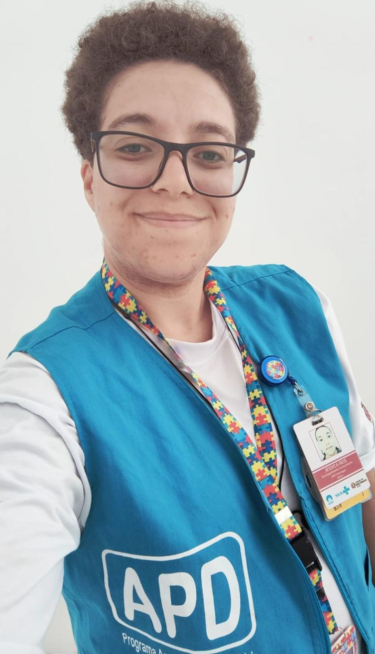 A imagem mostra uma pessoa sorrindo, usando óculos de armação preta e colete azul com a inscrição "APD - Programa Acompanhante de Pessoas com Deficiência". O colete azul tem o logotipo do programa estampado na frente.  A pessoa também usa um crachá de identificação com foto e uma fita colorida com estampa de peças de quebra-cabeça, símbolo frequentemente associado à conscientização sobre o autismo.