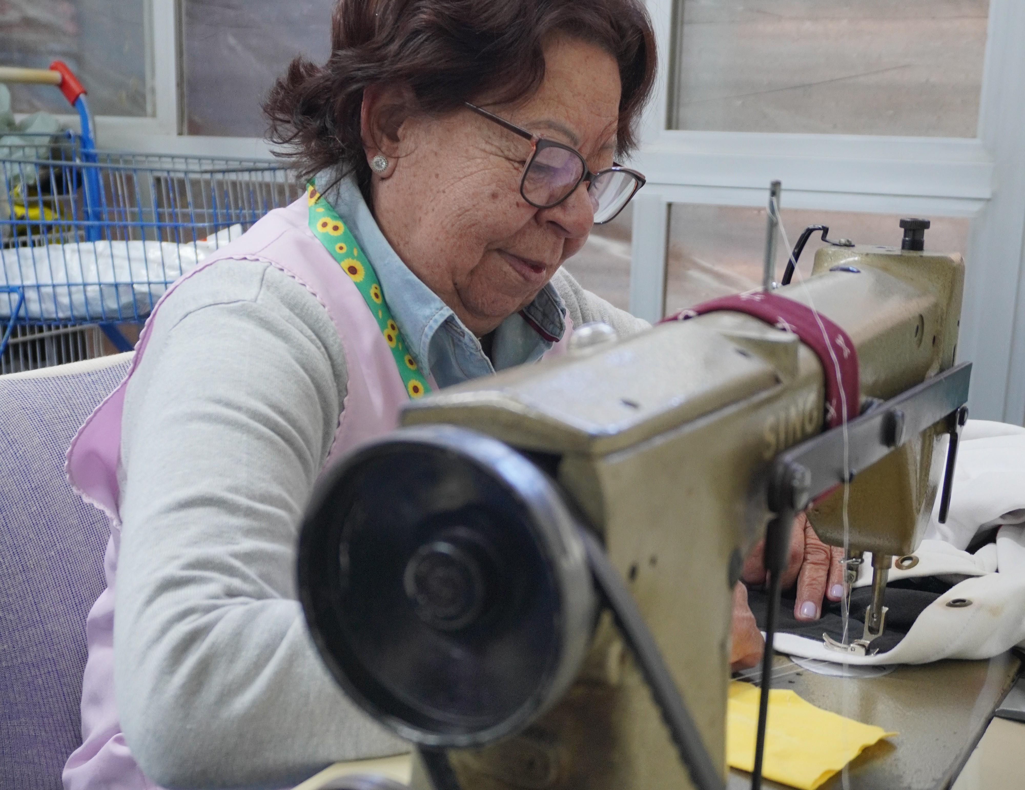 Foto em close de Maria costurando numa máquina. Ela tem pele clara a morena-clara, cabelos curtos castanhos escuros com fios grisalhos, levemente ondulados, e usa óculos de armação metálica. Veste camisa jeans clara sob um suéter cinza e um avental rosa; as alças do avental são verdes com estampa de flores amarelas. Usa pequenos brincos prateados.