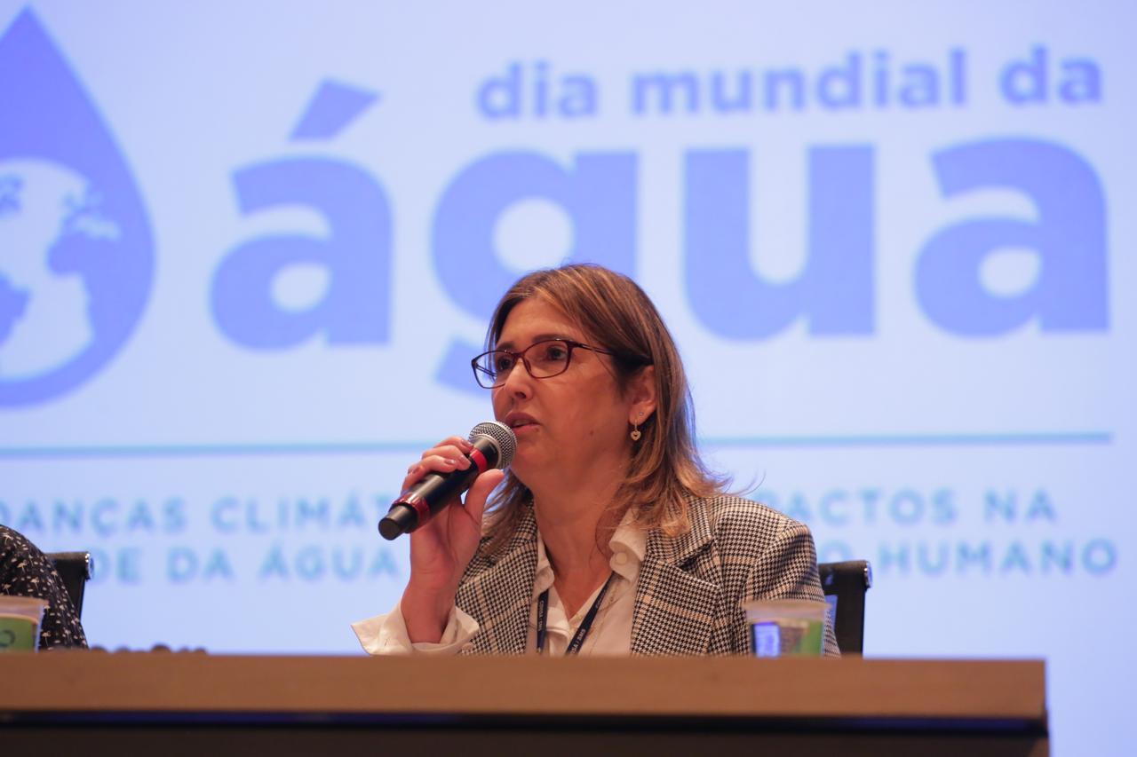 A foto mostra um fundo azul destacando o tema do evento Seminário da Água. Na mesa segurando um microfone está a organizadora do evento, a Diretora da Divisão Ambiental da Secretaria Municipal da Saúde, Magali Batistaali 