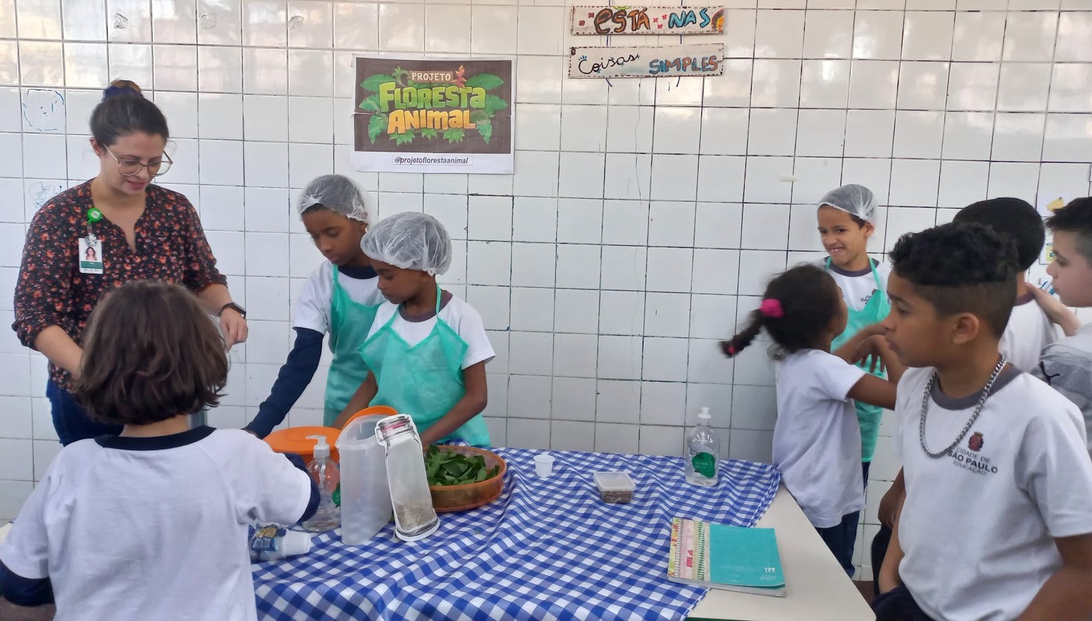 A imagem mostra uma atividade educativa envolvendo crianças em uma sala azulejada, com uma mesa coberta por toalha xadrez azul e branca; à esquerda, uma profissional com crachá acompanha os alunos na atividade. Duas crianças, usando toucas e aventais verdes, estão próximas à mesa, lidando com alimentos ou ingredientes — há uma vasilha com folhas verdes, potes transparentes e utensílios sobre a mesa. Outras crianças, algumas em fila, observam e participam da dinâmica, demonstrando curiosidade e interação. Na parede, há um cartaz com o título "Projeto Floresta Animal" e uma placa artesanal que diz: “Está nas coisas simples”. A cena transmite um ambiente de aprendizagem prática e coletiva, possivelmente relacionado à alimentação saudável, sustentabilidade ou aproveitamento dos alimentos, com foco em educação ambiental e hábitos saudáveis.