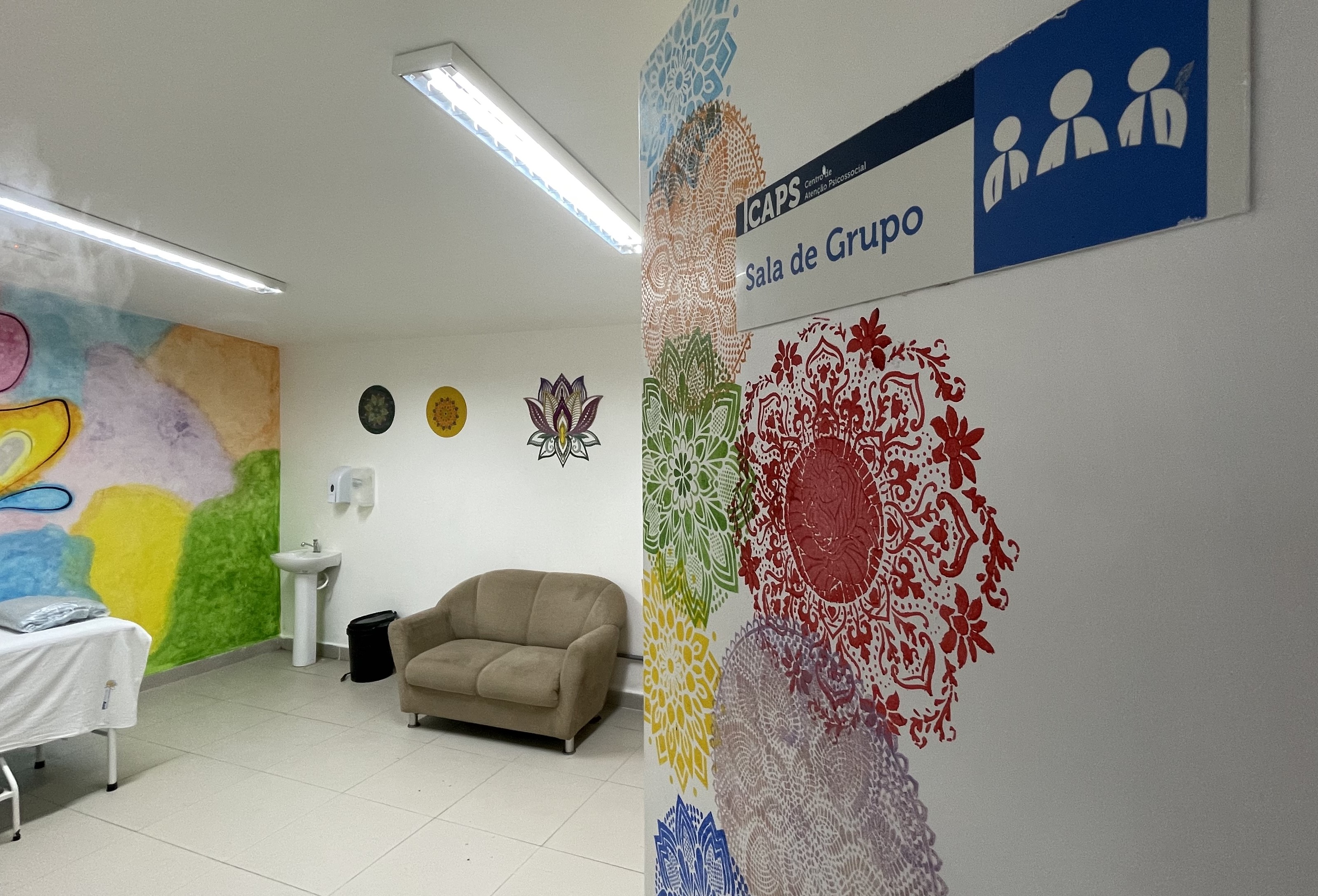 A imagem mostra o interior de uma sala identificada como “Sala de Grupo”, pertencente a um CAPS (Centro de Atenção Psicossocial). No canto direito, próximo à porta, há um mural colorido com mandalas de diferentes cores (vermelho, verde, amarelo, roxo e azul) pintadas sobre a parede branca. Mais ao fundo, vê-se uma parede decorada com desenhos de flores e formas geométricas, além de uma pintura artística com cores vivas (verde, amarelo, laranja, rosa e lilás) que cobre parte do canto esquerdo.  Dentro da sala, há um sofá bege de dois lugares, uma pia branca com dispenser de sabão fixado na parede e um cesto de lixo preto logo abaixo. À esquerda, aparece parcialmente uma maca coberta por lençóis. O ambiente é iluminado por luminárias retangulares de luz fria no teto, transmitindo uma atmosfera limpa e organizada, com decoração voltada para transmitir acolhimento e tranquilidade.       