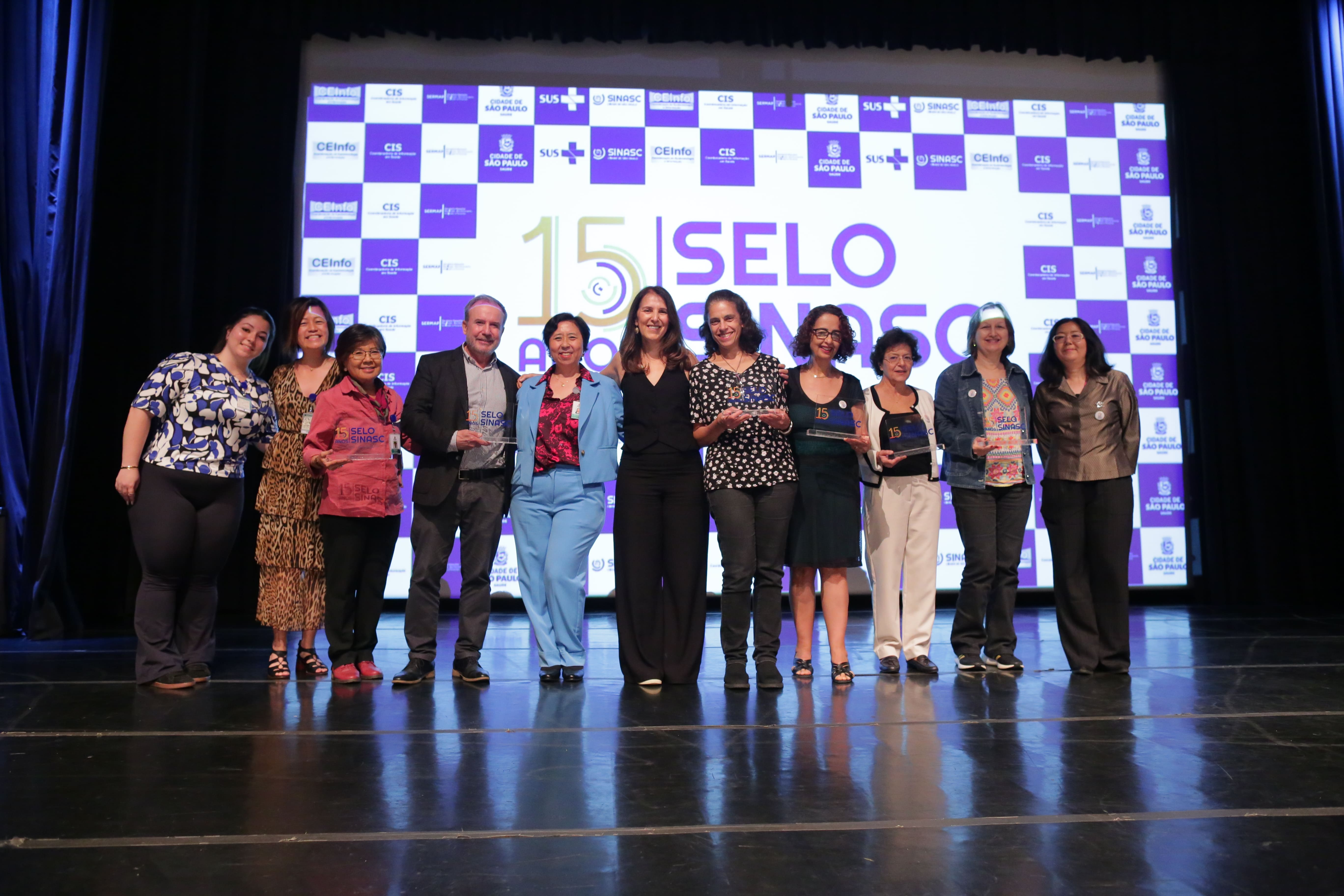 A imagem mostra um grupo de pessoas posando juntas em um palco durante um evento de premiação. Elas sorriem para a câmera, segurando troféus que parecem ser prêmios de reconhecimento. Ao fundo, há um grande telão com a marca do evento, destacando as palavras "15 anos Selo SINASC" junto com logotipos e marcas institucionais relacionadas ao SUS (Sistema Único de Saúde) e ao município de São Paulo. O grupo é composto por homens e mulheres, com uma diversidade de idades