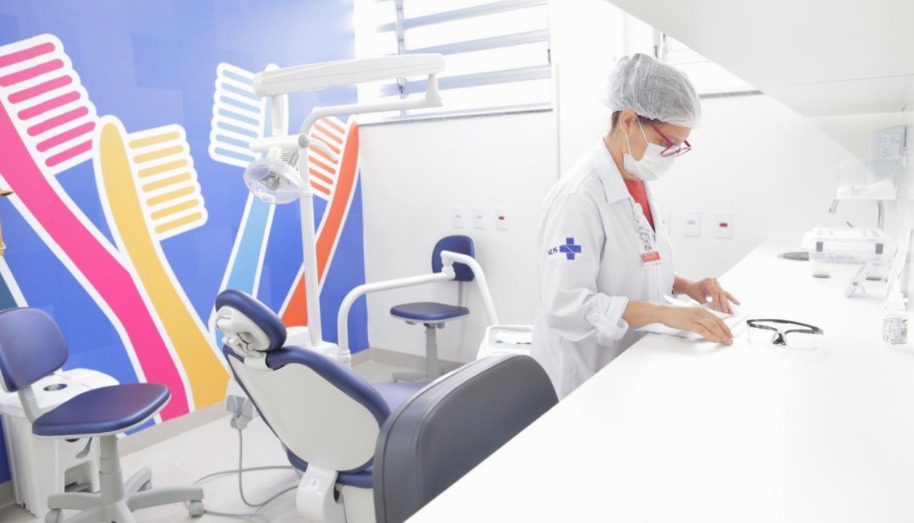 A foto mostra uma profissional de saúde em uma sala na qual se vê uma cadeira de dentista e uma ilustração de escovas de dentes coloridas adesivada na parede