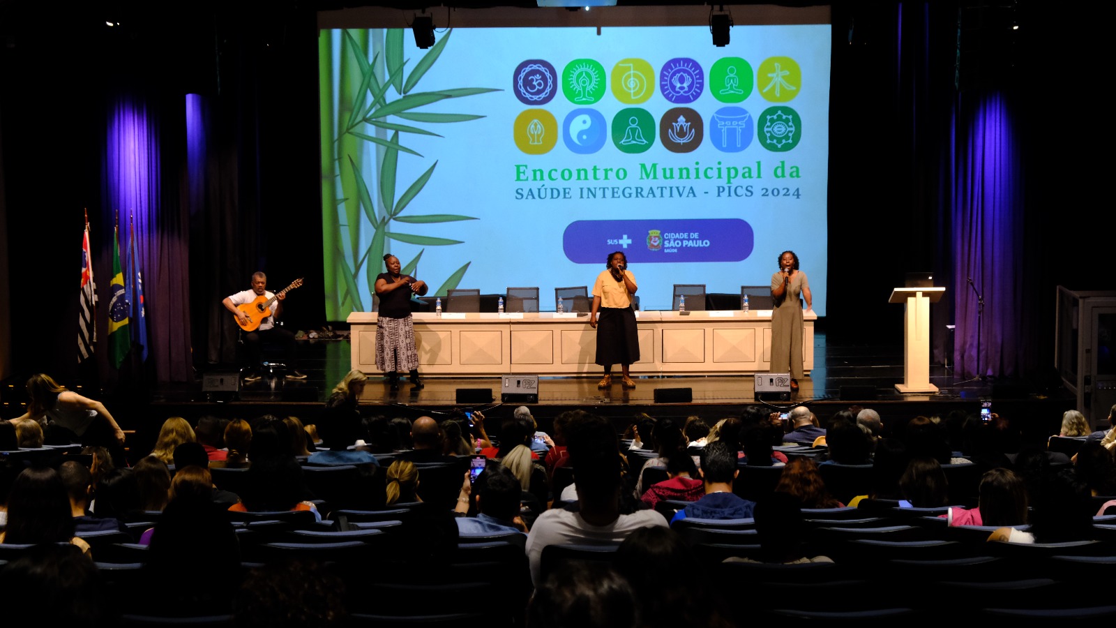 A foto mostra a apresentação musical de três mulheres negras e músico com violão 