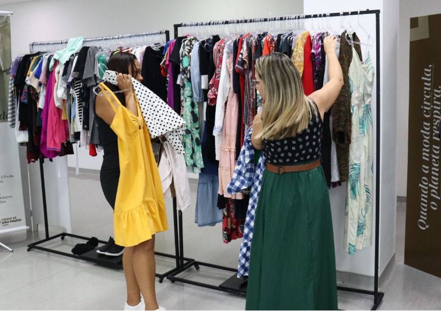 A foto mostra duas mulheres, uma de costas e outra de perfil, mexendo em uma arara com roupas; uma delas segura um vestido amarelo à frente do corpo 