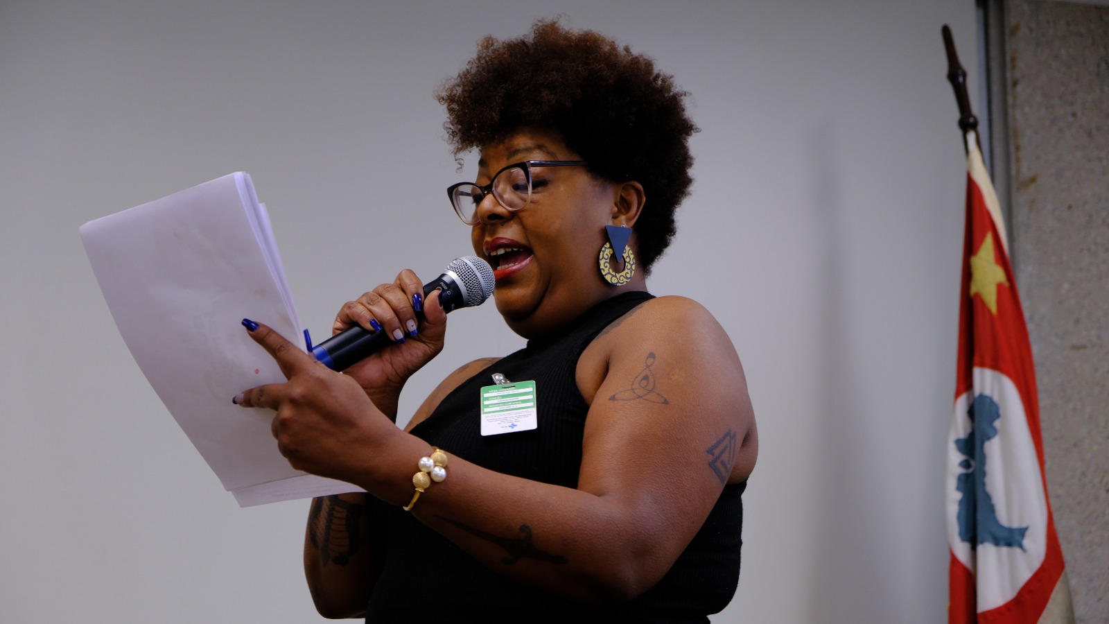 A imagem mostra Ana Cecília, uma mulher negra discursando enquanto segura um microfone e papéis. Ela usa óculos, um vestido preto, brincos grandes e coloridos, além de exibir tatuagens no braço. Ao fundo, à direita, há uma bandeira parcialmente visível com uma estrela amarela em destaque.