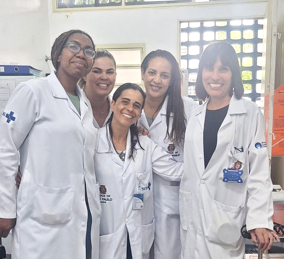 A imagem mostra cinco mulheres sorrindo, vestindo jalecos brancos, que indicam um ambiente profissional de saúde. Todas estão reunidas em um espaço interno bem iluminado, com paredes claras e detalhes típicos de uma sala de trabalho, como prateleiras, documentos e janelas com grade. Os jalecos apresentam logotipos do SUS (Sistema Único de Saúde). Vanessa está a direita, com cabelos pretos médios e franja.