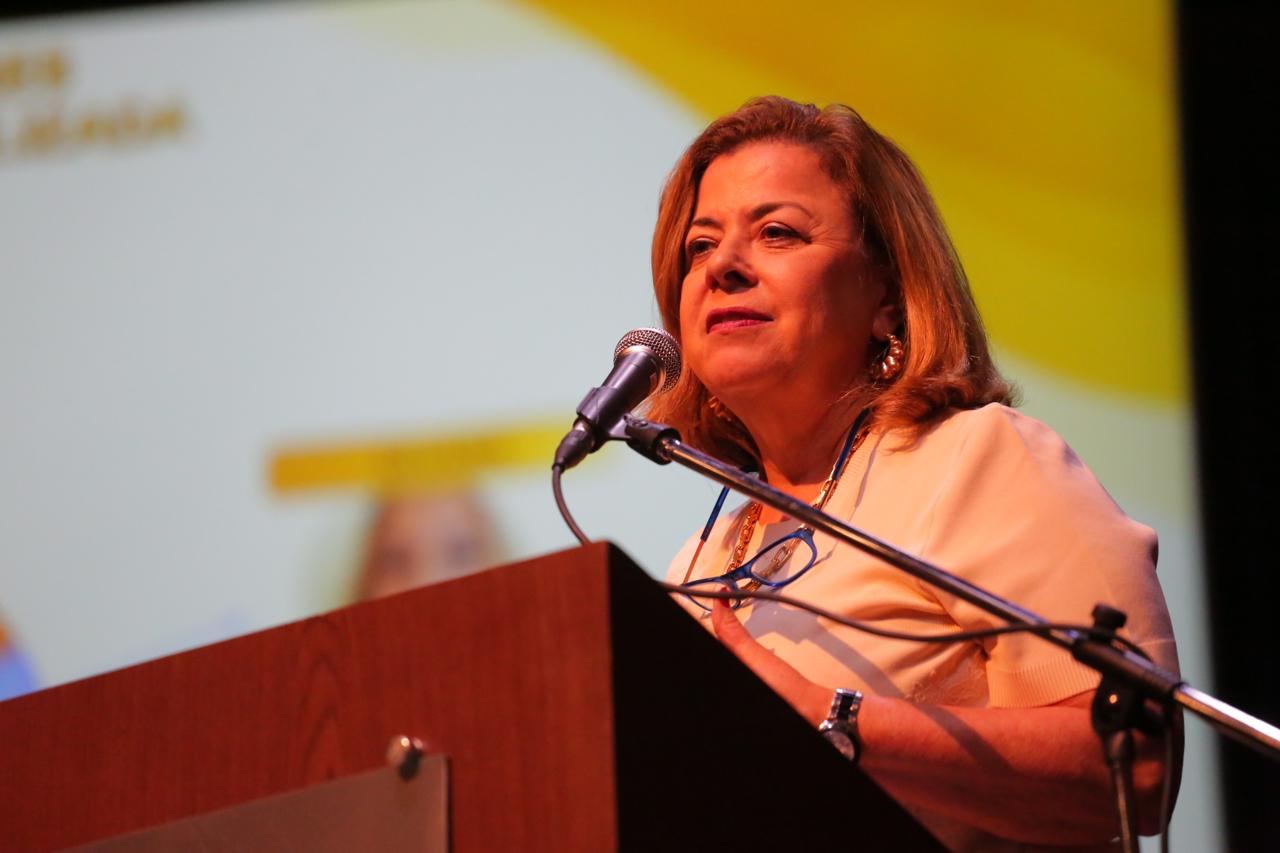 A imagem mostra uma mulher falando em um evento. Ela está atrás de um púlpito de madeira, com um microfone ajustado à sua altura. Ela veste uma blusa clara, usa colares e brincos dourados e tem um relógio no pulso esquerdo. Seus óculos estão pendurados no pescoço. Ao fundo, há uma tela de projeção com um design em tons amarelos e brancos