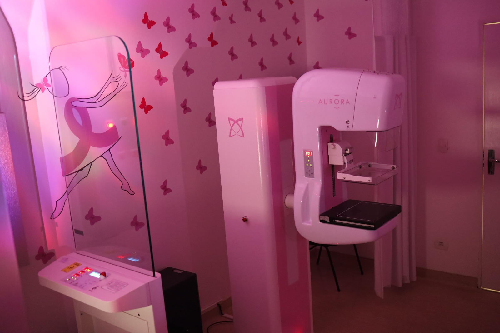 A imagem mostra uma sala equipada com um mamógrafo da marca "Aurora", utilizado para exames de mamografia. O ambiente tem uma iluminação suave em tons de rosa e decoração com borboletas e uma ilustração de uma mulher estilizada, incorporando o laço rosa, símbolo da campanha de conscientização sobre o câncer de mama