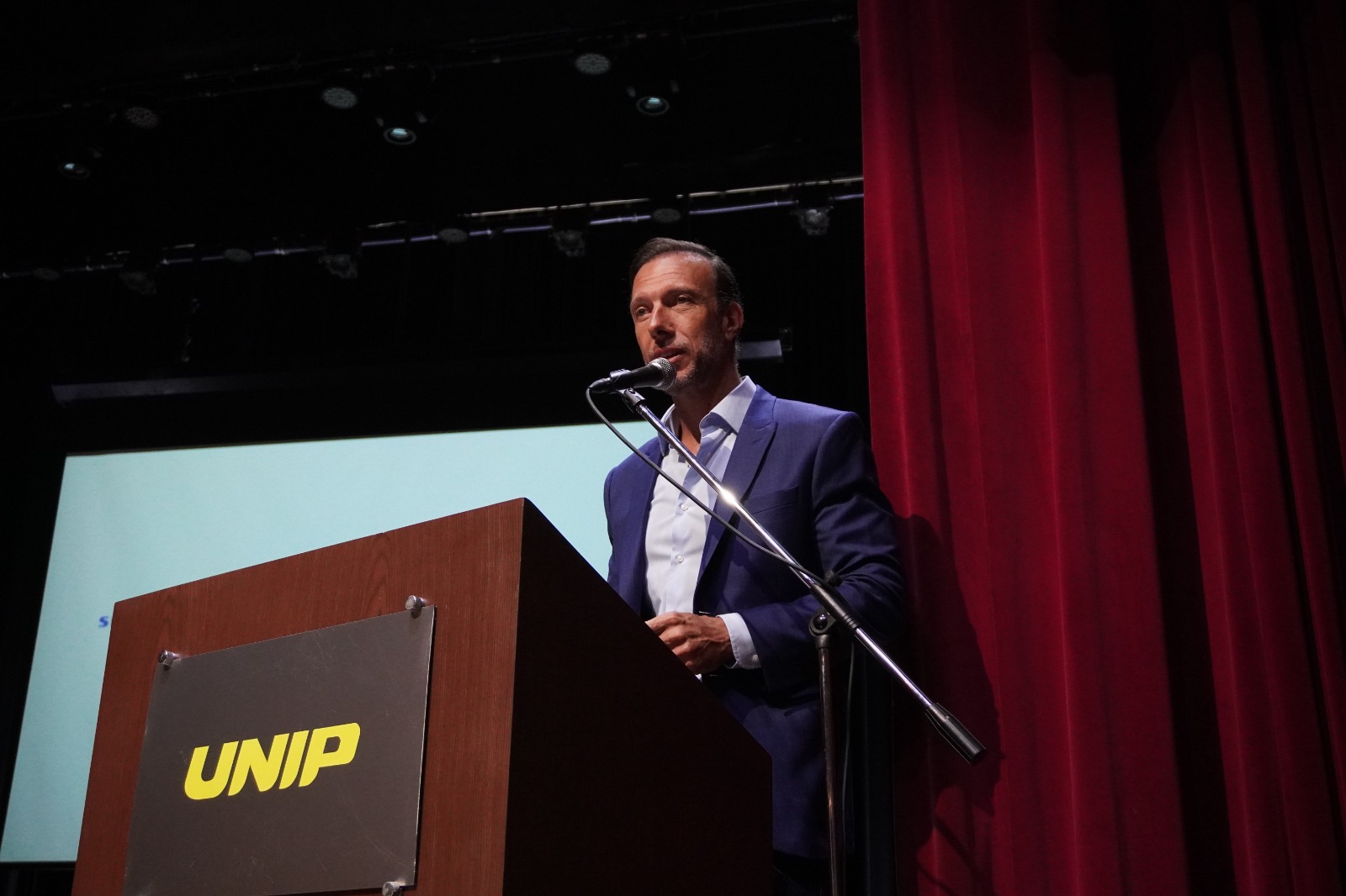 A imagem mostra um homem de terno azul e camisa branca discursando em um evento. Ele está em um palco, posicionado atrás de um púlpito de madeira com a logo da UNIP (Universidade Paulista). O microfone está fixado em um suporte, e ele aparenta estar focado em sua fala. Ao fundo, há uma tela de projeção parcialmente visível, além de cortinas vermelhas que compõem o cenário do auditório.