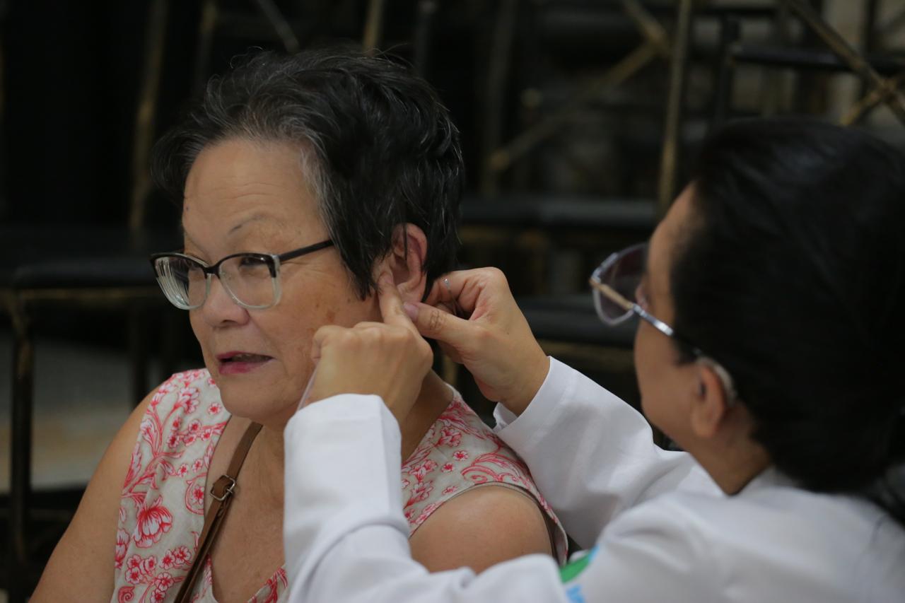 A imagem mostra uma profissional de saúde, vestindo um jaleco branco, realizando um procedimento na orelha de uma mulher idosa de óculos e cabelo curto. A paciente está vestindo uma blusa estampada e parece atenta ao que está sendo feito. A profissional segura delicadamente a orelha da paciente, possivelmente aplicando acupuntura auricular ou colocando um acessório terapêutico. O ambiente sugere um espaço de atendimento coletivo, com cadeiras ao fundo.
