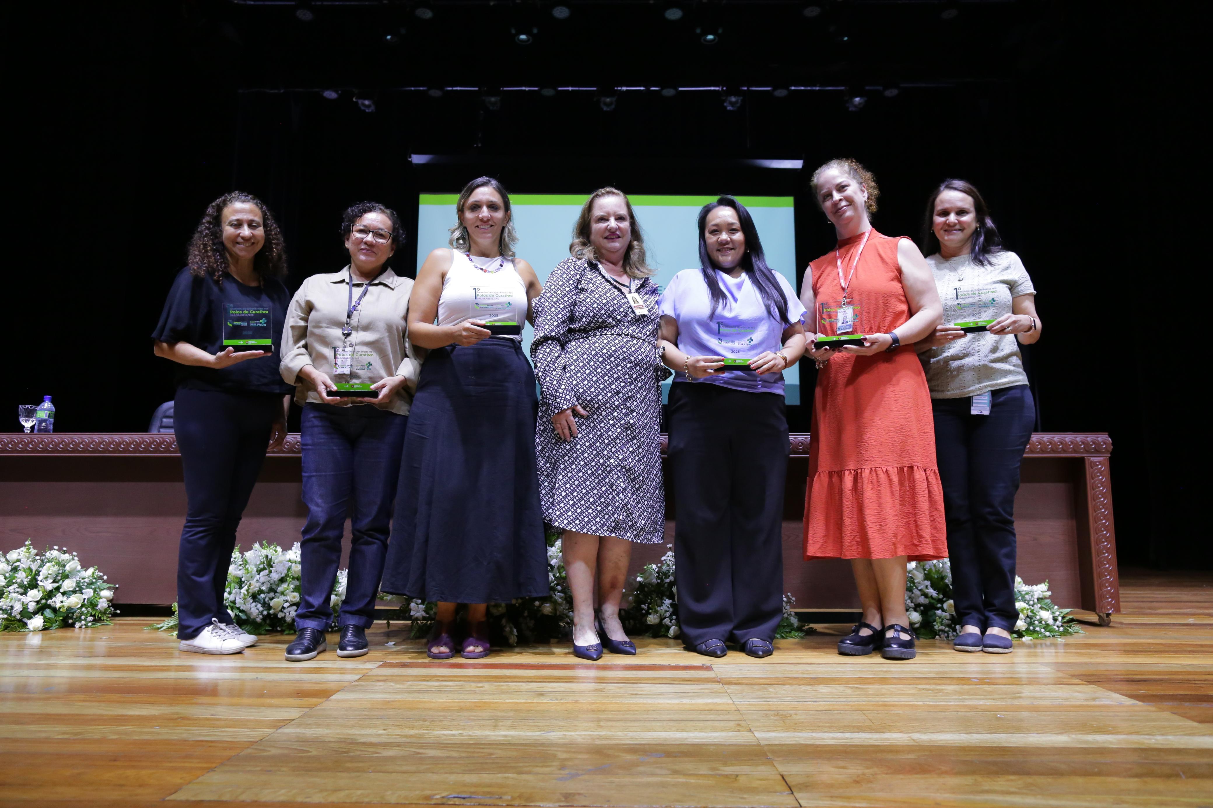 A imagem mostra um grupo de sete mulheres em um palco, posando para uma foto durante um evento de premiação. Todas seguram troféus ou placas de reconhecimento, indicando que foram homenageadas. Elas vestem roupas formais ou casuais elegantes e sorriem para a câmera. O cenário inclui um telão ao fundo, uma mesa decorada com flores e um ambiente que sugere um evento institucional ou acadêmico. A iluminação destaca o palco, criando uma atmosfera solene e comemorativa