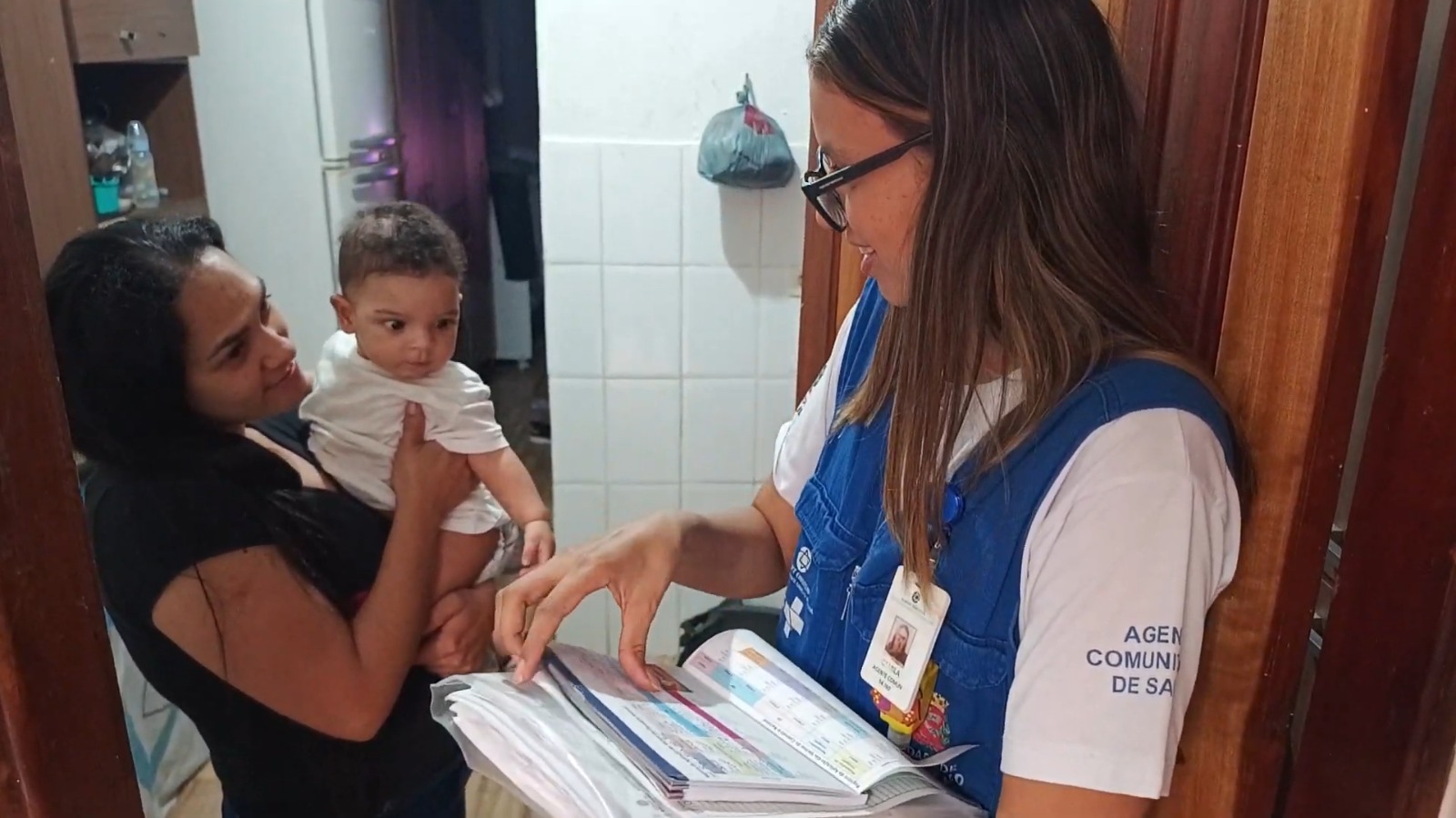 A imagem mostra uma cena interna, onde três pessoas estão interagindo em uma residência. À direita, está a agente comunitária de saúde, a mesma mulher da imagem anterior, que usa camiseta branca e colete azul da Prefeitura de São Paulo, com seus cabelos longos, lisos e castanhos, e óculos pretos. Ela segura uma pasta com documentos e parece estar explicando ou orientando sobre algo contido nesses materiais.  À esquerda, há uma mulher com cabelos longos, pretos e pele morena, vestida com uma camiseta preta, que segura um bebê no colo. Ela olha atentamente para a agente, com expressão de interesse e atenção. O bebê tem cabelos curtos e cacheados, pele morena clara e usa uma roupinha branca. Ele está curioso, olhando fixamente na direção da pasta da agente.