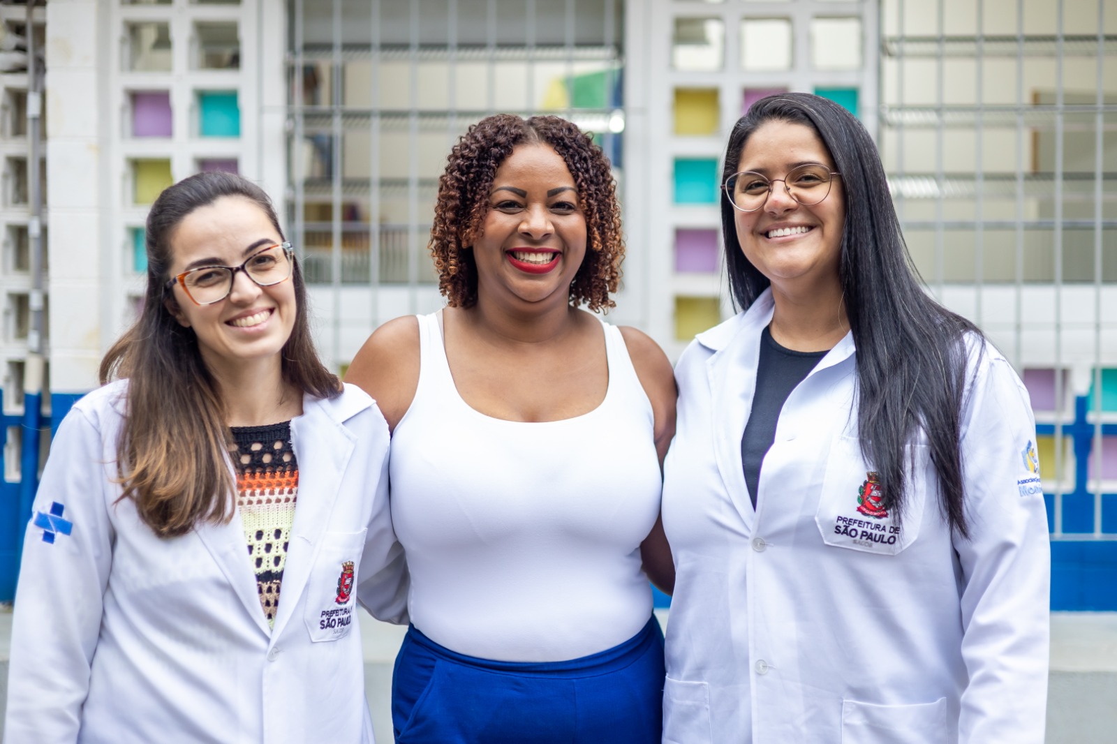 Com certeza! Aqui está uma descrição detalhada da imagem:  A imagem apresenta um grupo de três mulheres sorrindo e posando juntas em frente a uma parede com janelas coloridas.  À esquerda está Lucy, uma mulher de pele clara com cabelos longos e castanhos lisos, divididos ao lado, usa óculos de aro escuro. Ela veste um jaleco branco sobre uma blusa estampada com padrões geométricos em tons de laranja, preto e branco. No jaleco, do lado esquerdo do peito, há um símbolo azul que se assemelha a uma cruz dentro de um círculo. Do lado direito, há um brasão com o texto "PREFEITURA DE SÃO PAULO".  No centro está Camila, uma mulher negra e cabelos castanhos cacheados e volumosos. Ela está sorrindo abertamente, mostrando os dentes. Ela veste uma regata branca e uma calça ou saia azul royal de cintura alta. Seus lábios estão pintados de vermelho vivo.  À direita está Ana Caludia, uma mulher de pele clara com cabelos longos, lisos e pretos. Ela usa óculos de aro claro e veste um jaleco branco sobre uma blusa preta. No jaleco, do lado direito do peito, há o mesmo brasão com o texto "PREFEITURA DE SÃO PAULO" visto na mulher à esquerda.