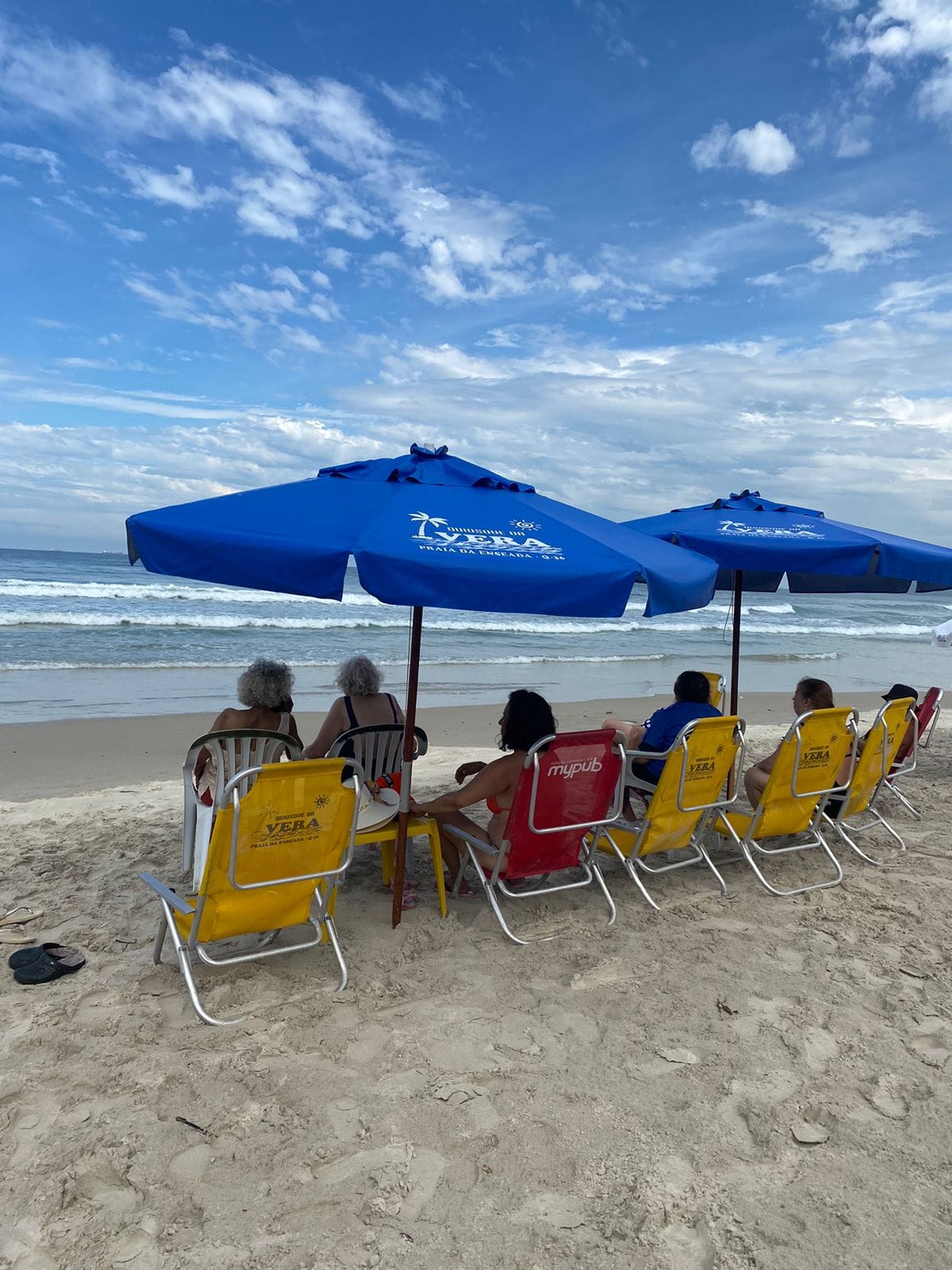 A imagem mostra um grupo de pessoas idosas sentadas em cadeiras de praia voltadas para o mar, sob a sombra de grandes guarda-sóis azuis com a inscrição “Vera Praia da Enseada - G.J.E.”. A cena ocorre em uma praia de areia clara e mar calmo, sob um céu parcialmente nublado com algumas aberturas de azul, transmitindo um clima tranquilo.  As cadeiras, amarelas e vermelhas, estão dispostas lado a lado na faixa de areia, e os banhistas parecem relaxar e contemplar o mar, em um momento de descanso e convivência tranquila. Algumas pessoas conversam entre si, enquanto outras apenas apreciam a paisagem. Ao lado esquerdo da imagem, há pares de chinelos na areia, reforçando o tom de informalidade e conforto da situação.