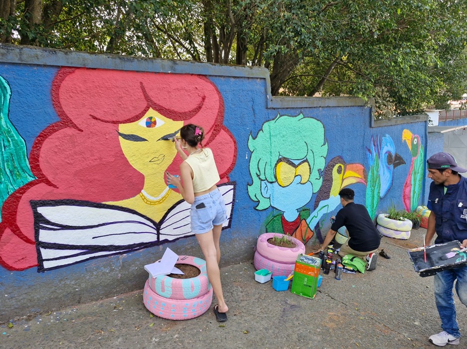 A imagem mostra uma ação de arte urbana em andamento, com foco na pintura de um mural colorido em um muro externo. Três pessoas estão envolvidas na atividade: uma jovem pinta os detalhes do rosto de um personagem feminino de cabelos vermelhos, enquanto outra pessoa trabalha nos contornos de um personagem azul com cabelo verde e óculos escuros. Um terceiro indivíduo caminha próximo, segurando uma bandeja com tinta.  O mural apresenta personagens estilizados com forte apelo visual: à esquerda, uma figura feminina com um livro aberto, olhos fechados e um símbolo colorido na testa (lembrando o símbolo dos Objetivos de Desenvolvimento Sustentável da ONU); ao centro, um personagem com pele azul, expressão séria e pássaros tropicais ao fundo, como tucanos e araras, reforçando a temática ambiental.