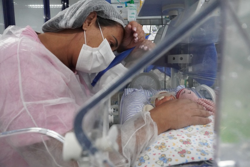 Na foto, uma mulher adulta está inclinada sobre uma incubadora de bebê. Ela usa uma touca cirúrgica cinza clara e uma máscara facial branca que cobre seu nariz e boca. Sua pele parece ter um tom moreno claro, e seus cabelos escuros estão completamente cobertos pela touca. Seus olhos estão voltados para o bebê dentro da incubadora. Uma de suas mãos está delicadamente posicionada sobre a lateral da incubadora, tocando a bebê, enquanto a outra parece estar perto de seu rosto. Ela veste uma espécie de camisola ou roupão hospitalar de cor rosa claro.  Dentro da incubadora, podemos ver um bebê muito pequeno, deitado de costas sobre um tecido estampado com desenhos infantis coloridos. O bebê parece estar envolto em um tecido claro e tem alguns tubos ou fios finos conectados ao seu corpo, sugerindo que ele está recebendo cuidados médicos. 