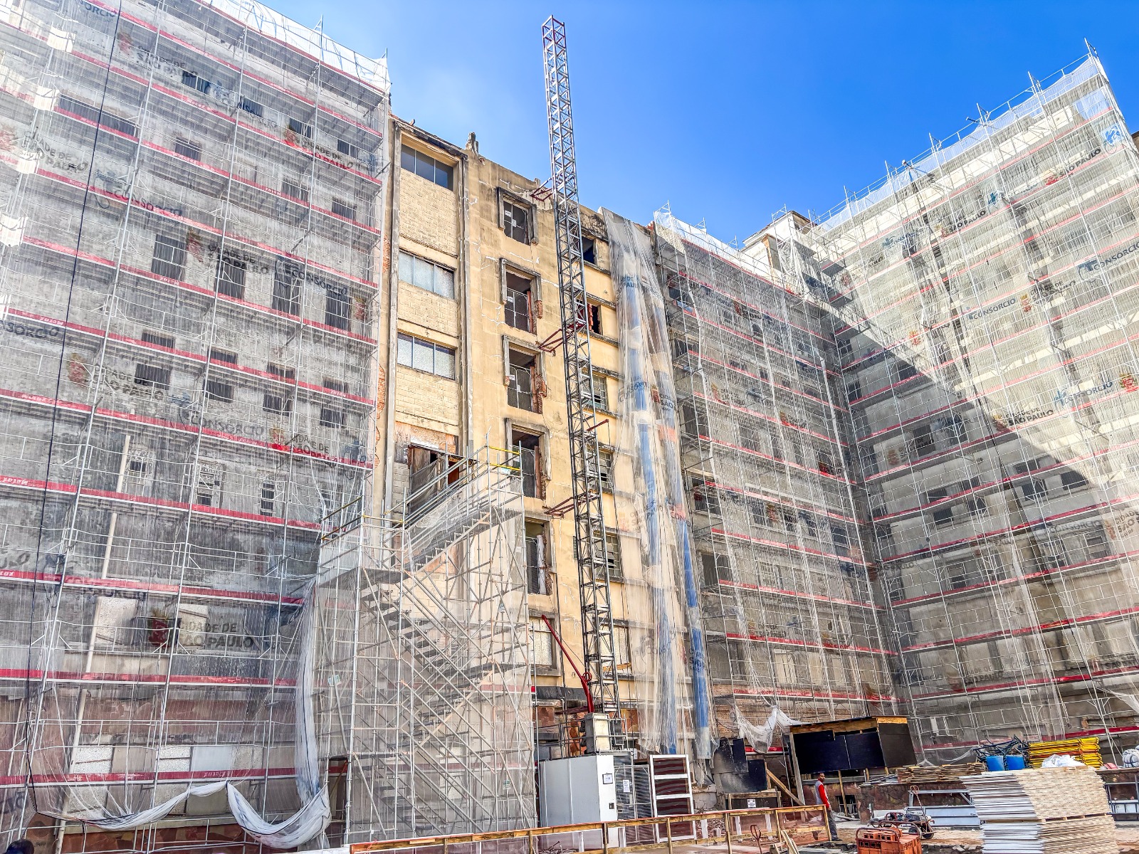 A imagem mostra um prédio de grande porte em processo de construção. O edifício tem formato em “U” e estrutura robusta, com andaimes e telas de proteção cobrindo quase toda a fachada. As telas são de cor clara com faixas horizontais vermelhas e marcas com os dizeres “CIDADE DE SÃO PAULO”.  A parte central do prédio está mais exposta, com a alvenaria e concreto aparentes, janelas quebradas ou sem vidros, e sinais de deterioração. Algumas paredes já foram descascadas, revelando tijolos. No centro da imagem, uma escada metálica provisória leva até os andares superiores, conectando aos andaimes. Também há uma estrutura vertical de guindaste ou elevador de carga.  Na base do edifício, há materiais de construção empilhados — como chapas de madeira compensada, vergalhões, bobinas de fios e blocos. Há também um contêiner branco e operários usando capacetes. O céu está limpo e azul, iluminando bem a obra. 
