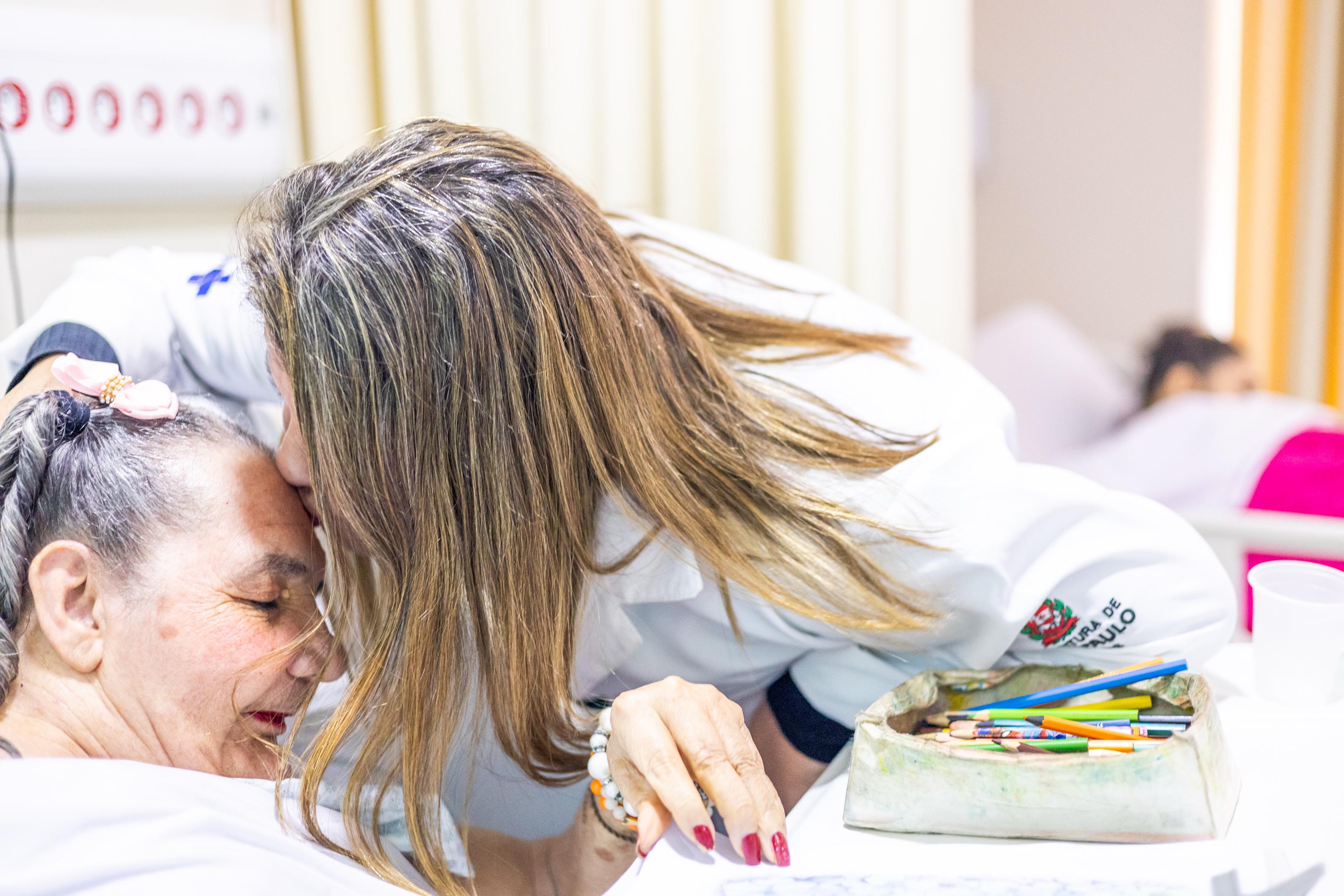 A imagem retrata um momento afetuoso dentro de um ambiente hospitalar. Shirley está vestida com jaleco branco com os logotipos do SUS e da Prefeitura de São Paulo, está inclinada e dá um beijo carinhoso na testa de uma paciente idosa. A profissional tem cabelos lisos, loiros com mechas claras, e unhas pintadas de vermelho. Ela segura delicadamente a mão da paciente, que tem pele clara, cabelos grisalhos presos em uma trança com um laço rosa e usa batom vermelho.  Sobre a mesa ao lado, há um recipiente com vários lápis de cor, sugerindo atividades lúdicas ou terapêuticas. Ao fundo, é possível ver outra paciente deitada em uma cama, parcialmente coberta por um cobertor rosa. O clima geral da imagem transmite acolhimento, cuidado e empatia no atendimento à pessoa idosa.