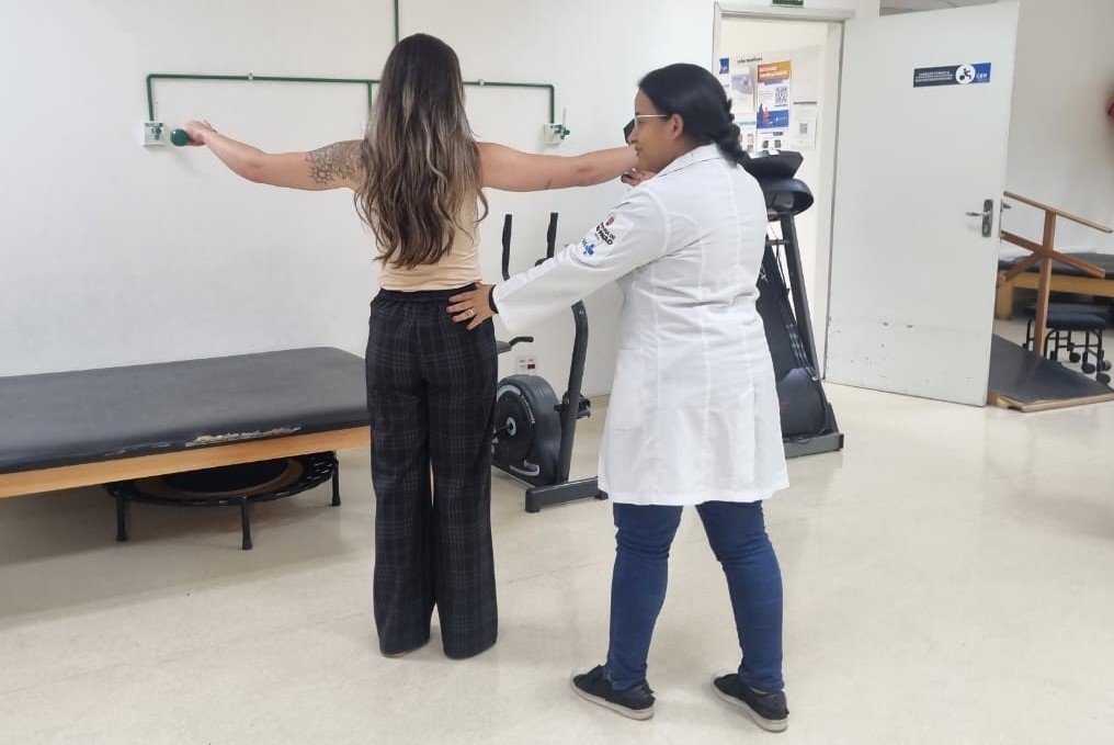 A imagem mostra um atendimento de fisioterapia em uma clínica ou unidade de saúde.  No centro, uma paciente de cabelos longos soltos está de pé, com os braços abertos em posição de exercício. Ela é acompanhada por uma profissional de saúde que veste jaleco branco com o logotipo do SUS na manga. A fisioterapeuta orienta e apoia a paciente, posicionando uma das mãos em suas costas e a outra no braço, garantindo correção postural durante a atividade.  O ambiente é equipado com aparelhos e recursos de reabilitação, como uma maca, bicicleta ergométrica, escada terapêutica e outros equipamentos ao fundo. A cena transmite um momento de reabilitação, cuidado especializado e acompanhamento próximo do profissional de saúde, reforçando a importância da fisioterapia no fortalecimento físico e na recuperação funcional.