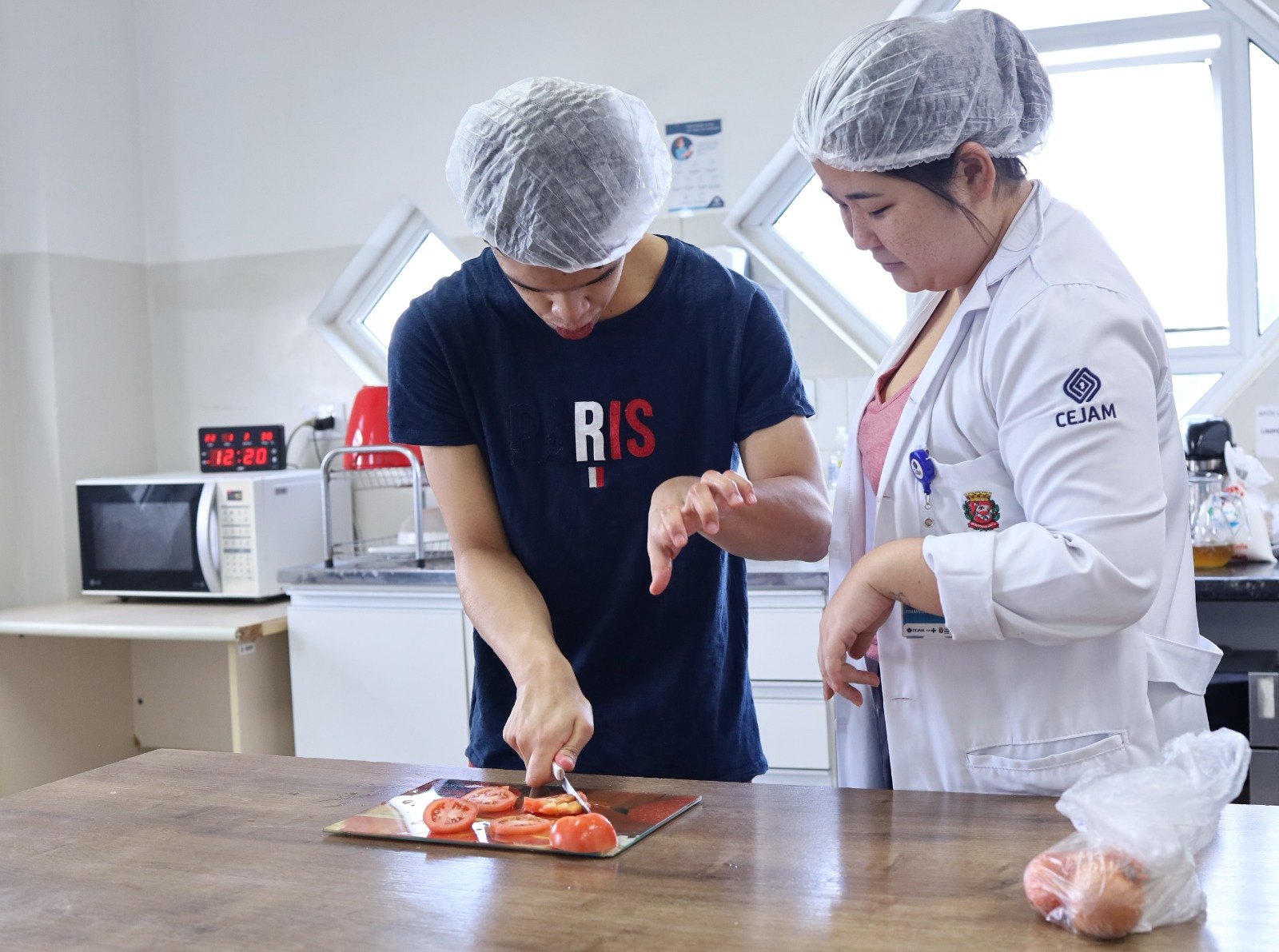 No centro, um jovem com touca descartável corta tomates em uma tábua, concentrado na tarefa. Ao lado dele, uma profissional de saúde ou educadora, também usando touca e jaleco branco com o logotipo do CEJAM e o brasão da Prefeitura de São Paulo, acompanha atentamente o processo, orientando e garantindo segurança.  Na bancada, além dos tomates cortados, há uma cebola dentro de um saco plástico. Ao fundo, é possível ver um micro-ondas, utensílios de cozinha e janelas que iluminam o ambiente.  A cena transmite ensino de autonomia, reabilitação e inclusão por meio de atividades do dia a dia, como o preparo de alimentos.