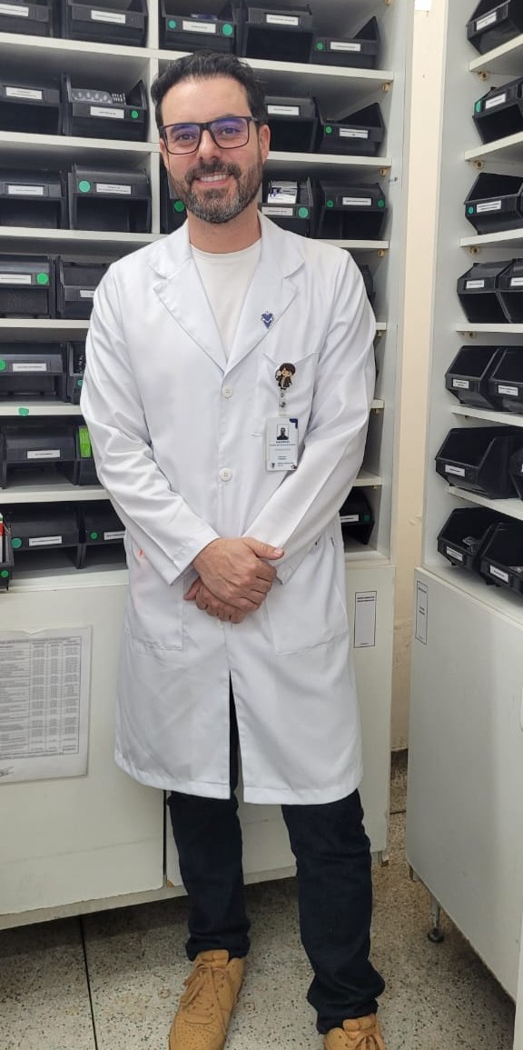 A imagem mostra um profissional da saúde em pé, posando para a câmera em um ambiente interno que aparenta ser uma farmácia ou almoxarifado de unidade de saúde. Ele veste um jaleco branco, usa óculos, tem barba curta e sorri de forma discreta, transmitindo uma postura acolhedora e confiante. As mãos estão cruzadas à frente do corpo.  No jaleco, há um crachá de identificação e um pequeno broche decorativo, indicando vínculo institucional. Ele usa calça escura e tênis de cor clara, em contraste com o jaleco.  Ao fundo, vêem-se prateleiras organizadas com caixas plásticas identificadas, típicas de armazenamento de medicamentos ou insumos, reforçando o contexto de trabalho na área da saúde. O ambiente é funcional, bem iluminado e organizado, característico de um espaço técnico-assistencial.