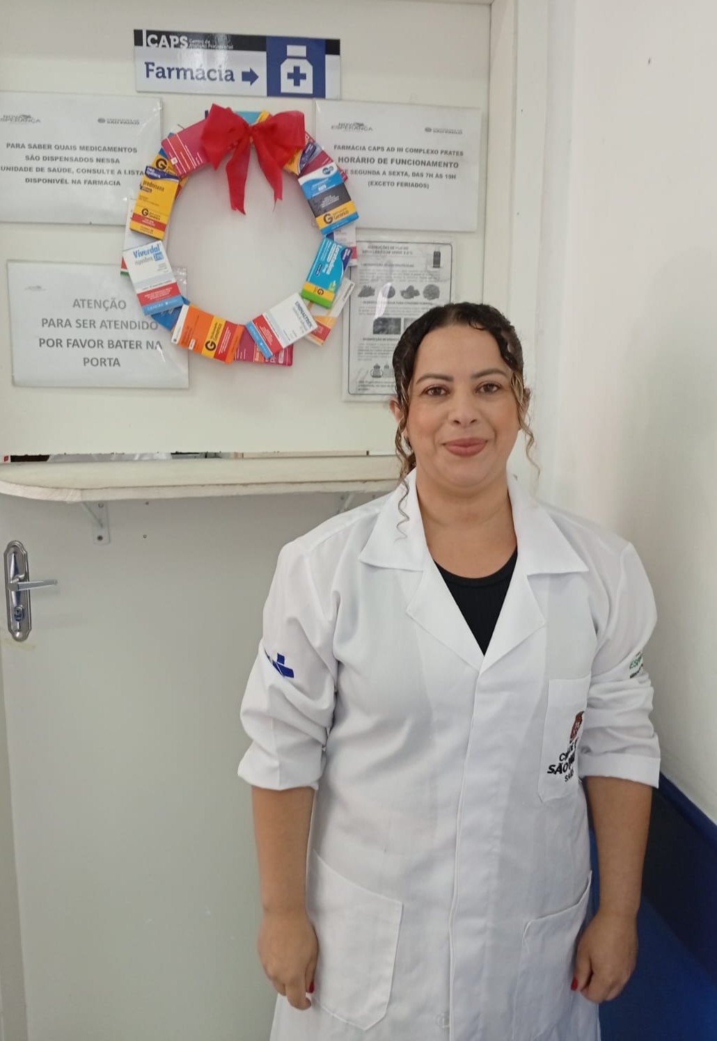 A imagem mostra uma profissional da saúde em pé, sorrindo levemente para a câmera, vestindo um jaleco branco, em um ambiente interno de serviço de saúde. Ela está posicionada em frente à porta de uma farmácia, indicada por placas fixadas na parede com a palavra “Farmácia” e orientações de atendimento e funcionamento.  Na porta, há uma guirlanda decorativa feita com caixinhas de medicamentos, dispostas em formato circular e finalizadas com um laço vermelho, o que sugere uma decoração temática, possivelmente relacionada a uma data comemorativa.  O ambiente é simples e funcional, com paredes claras e sinalização institucional, típico de uma unidade de saúde pública. A profissional aparece em postura relaxada, com os braços ao longo do corpo, transmitindo uma sensação de acolhimento e cuidado.