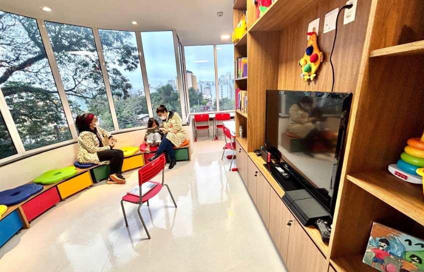 A imagem mostra um ambiente interno acolhedor e voltado ao atendimento infantil, possivelmente em um serviço de saúde ou espaço educativo.  À direita, há um móvel de madeira com prateleiras que abrigam brinquedos coloridos, livros e um televisor. O espaço é organizado e pensado para o entretenimento das crianças. No centro do ambiente, há cadeiras infantis vermelhas e bancos baixos com assentos coloridos encostados à parede.  À esquerda, próximo às janelas amplas, duas mulheres adultas e uma criança estão sentadas, interagindo de forma tranquila. As adultas usam máscaras e parecem conversar ou orientar a criança, que está sentada entre elas. As grandes janelas permitem a entrada de luz natural e oferecem vista para área arborizada e prédios ao fundo, sugerindo um local urbano.  O ambiente é bem iluminado, limpo e colorido, transmitindo uma sensação de cuidado, conforto e acolhimento, especialmente voltado ao público infantil.